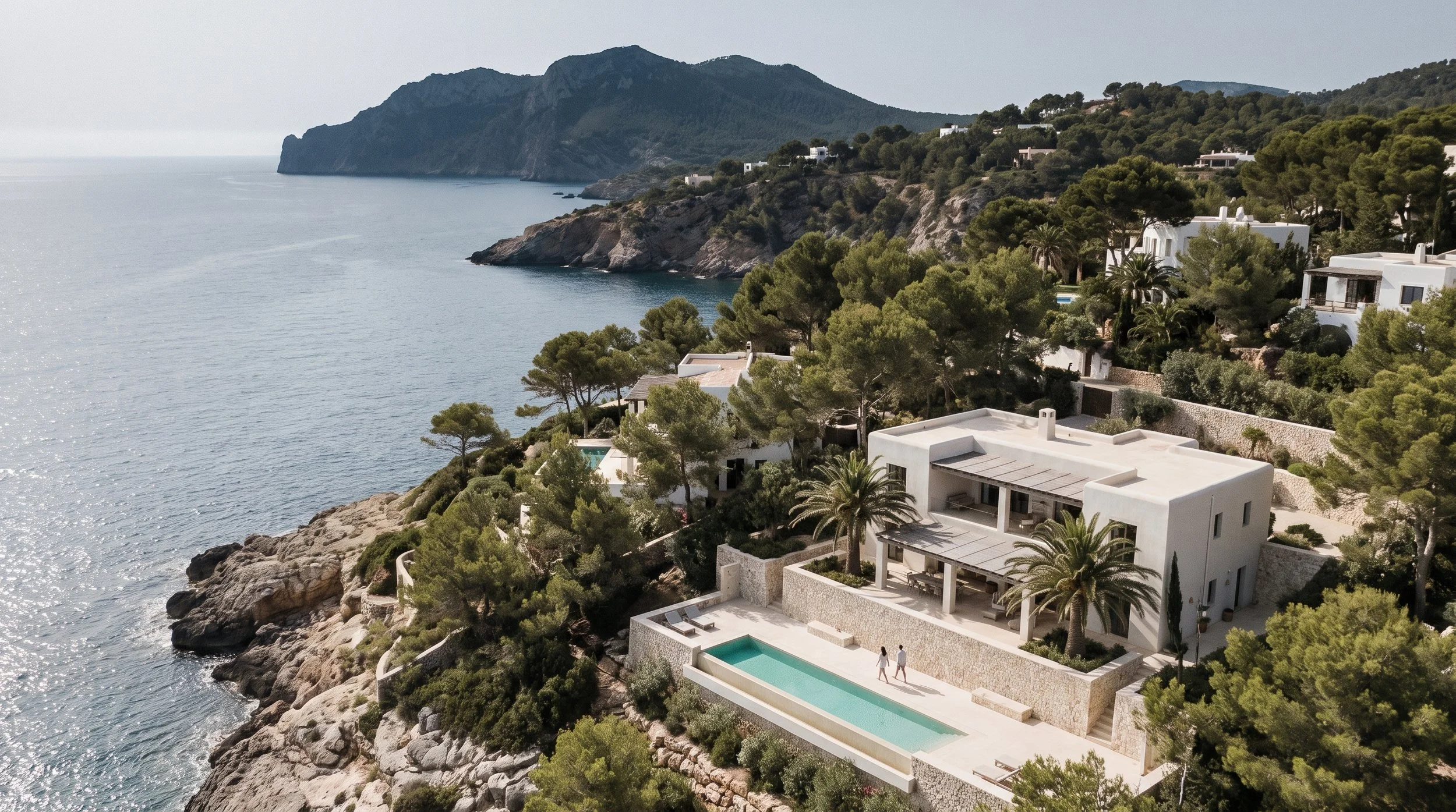 Aerial view of modern white Mediterranean-style villas on a cliff overlooking the ocean, with lush green trees and distant mountains in the background.