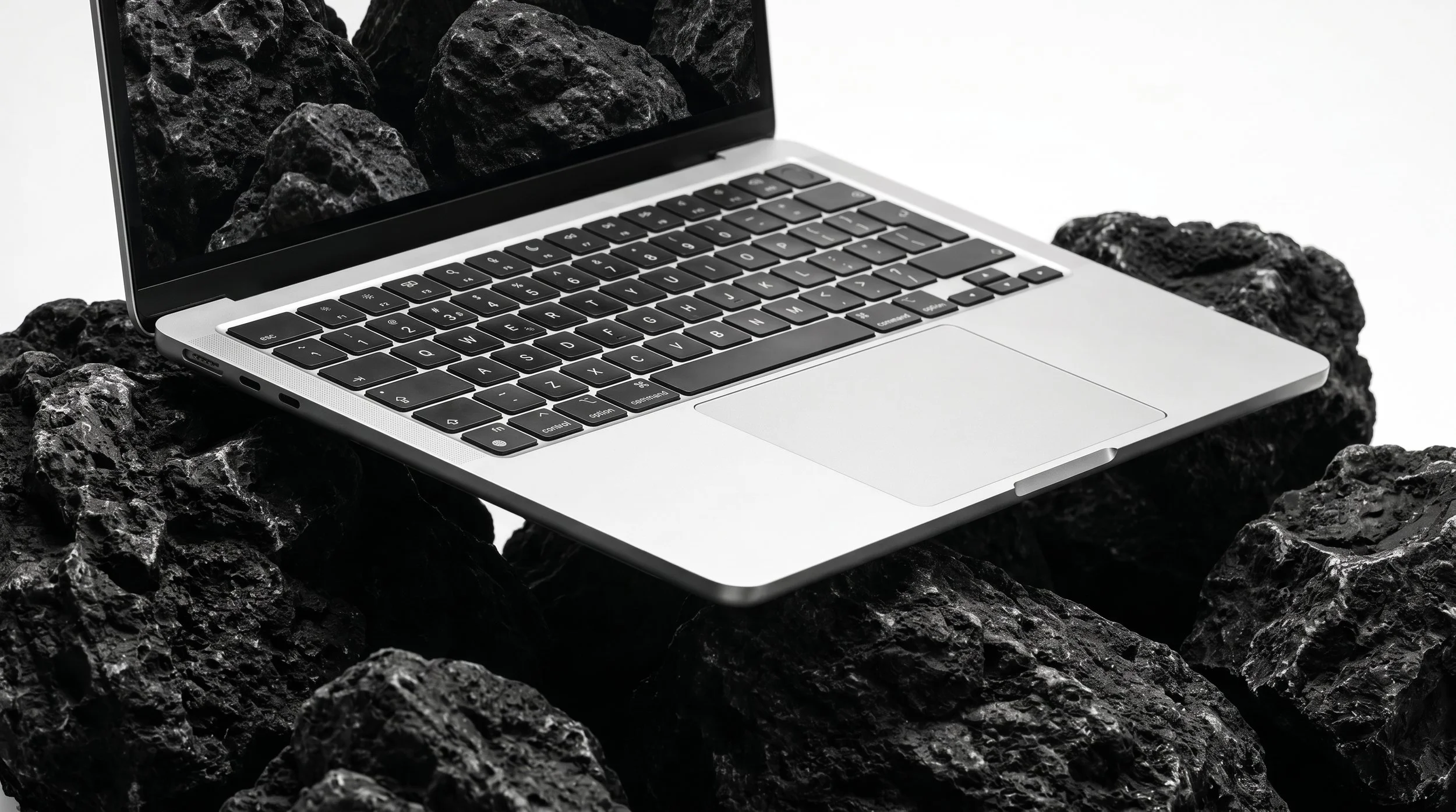 A modern laptop with black keyboard and silver body, sitting on black volcanic rocks with a plain white background.