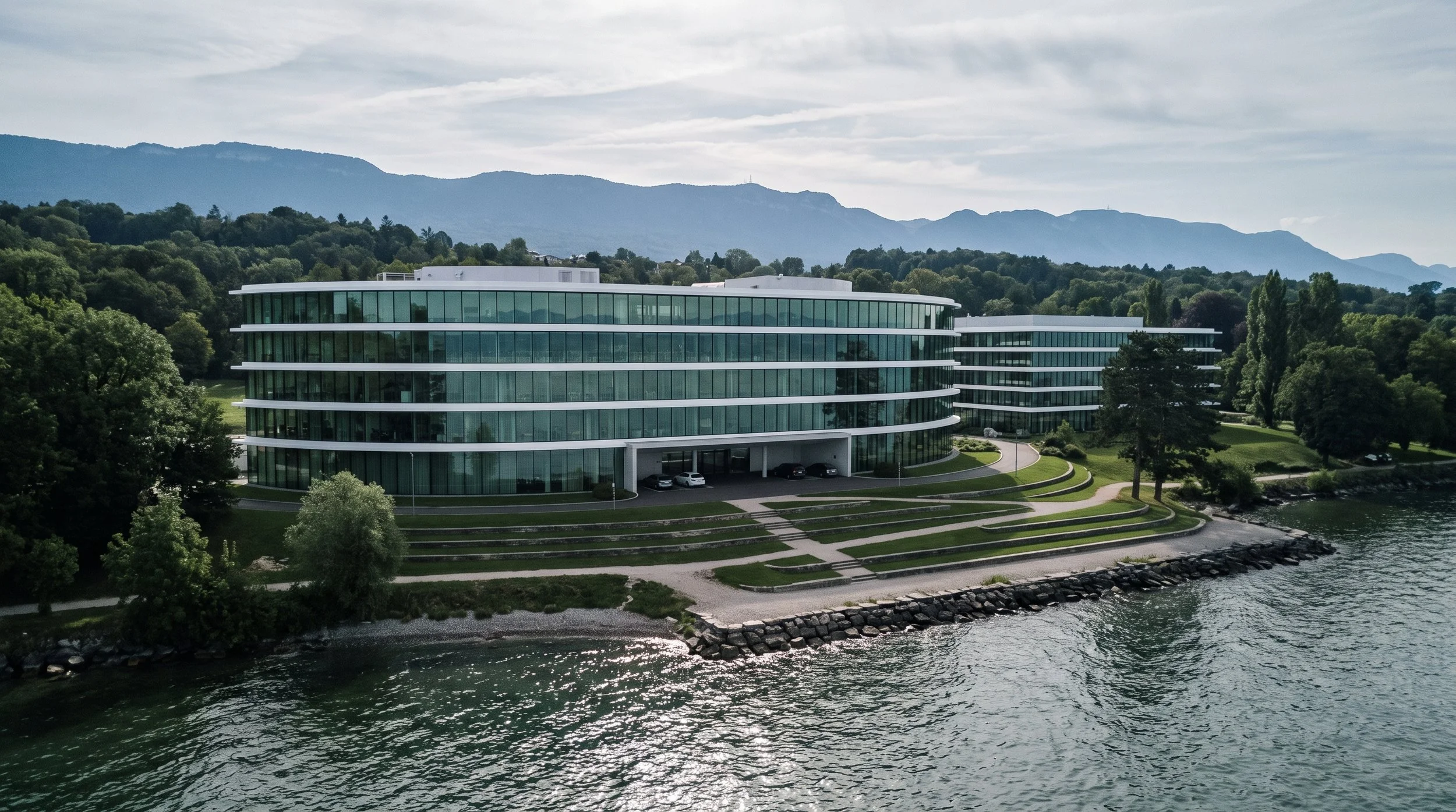 Modern glass office buildings situated on a hillside near a body of water, with mountains and a partly cloudy sky in the background.