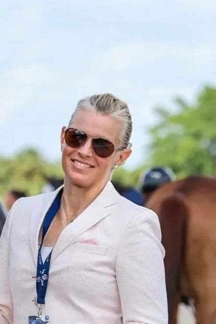 A woman with short gray hair wearing sunglasses, a white blazer, and a blue lanyard with a medal, standing outdoors with a horse and trees in the background.