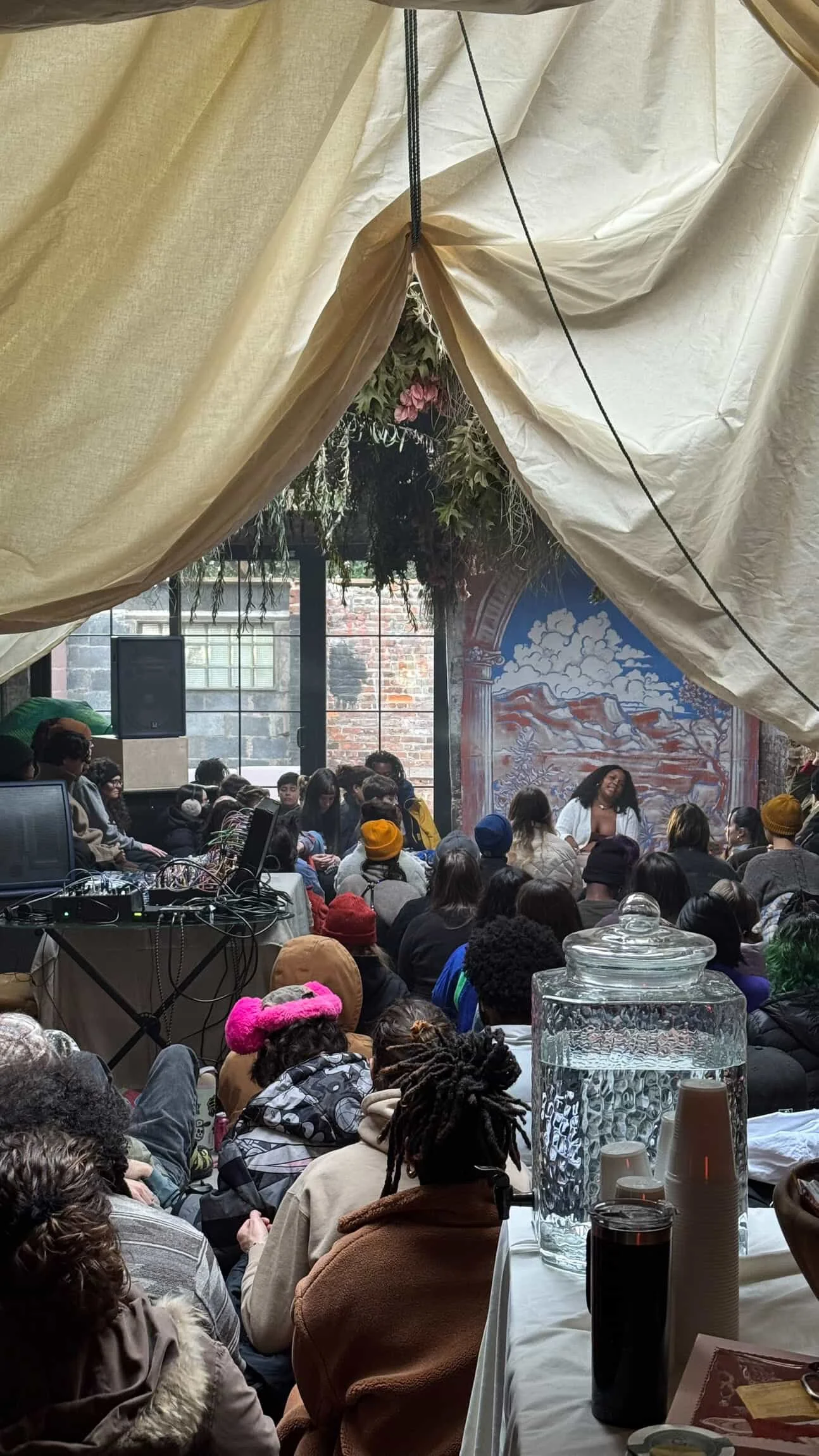 Large community gathering with Alexis Sāga speaking to a room of guests seated together before a colorful mural.