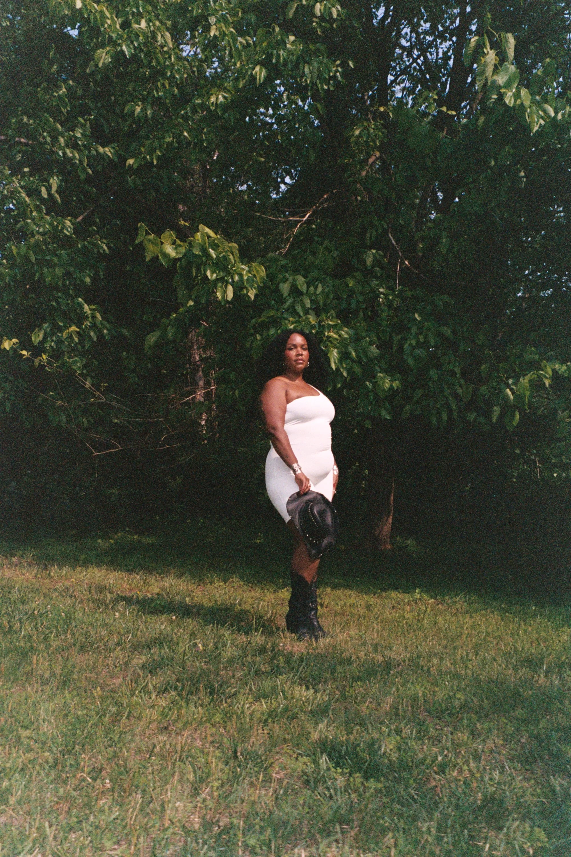 Alexis Sāga standing beneath a large leafy tree, dressed in white and holding a black hat.