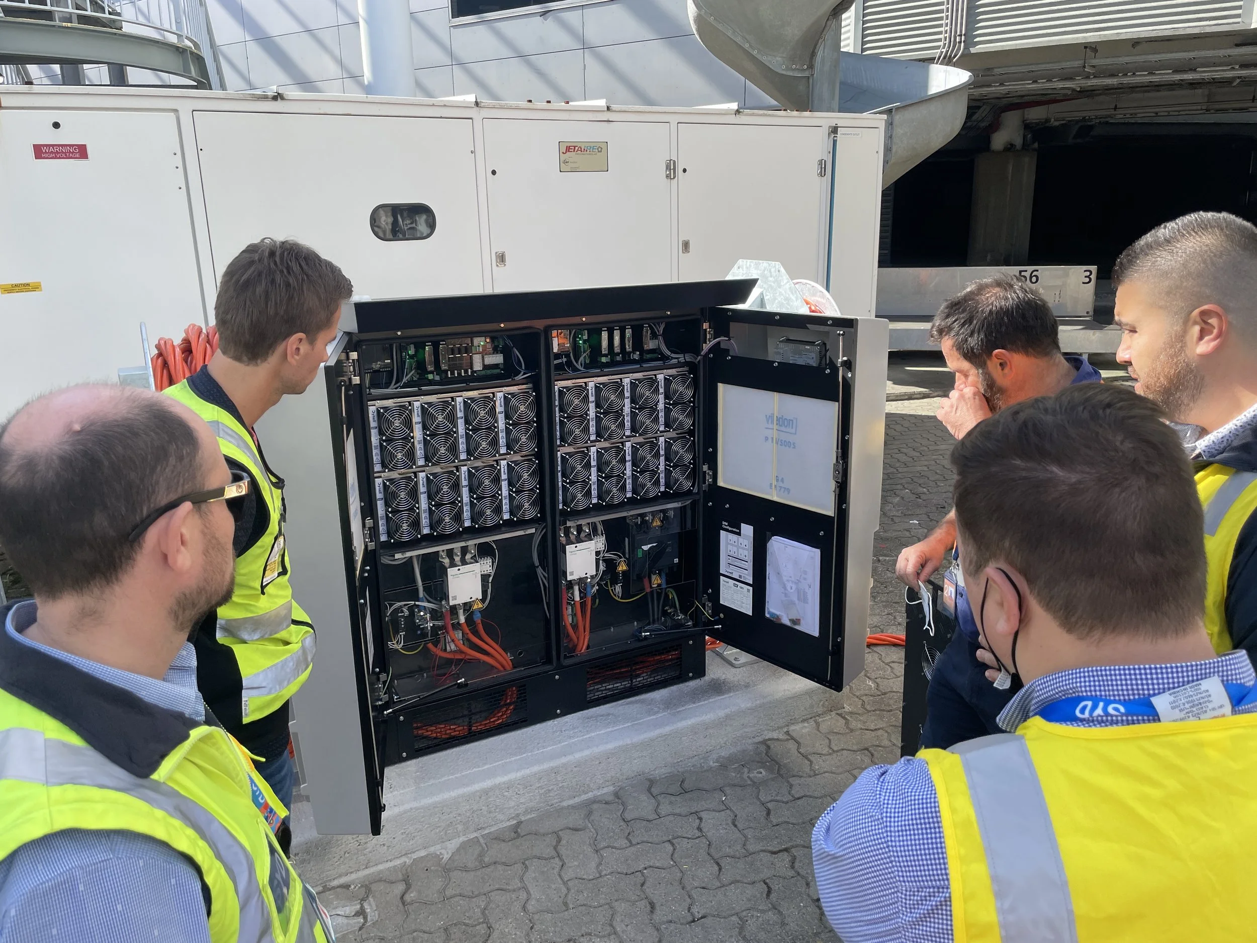 Group of Airport Equipment workers gathered around an open Dynell System, inspecting and discussing the components, wearing safety vests.