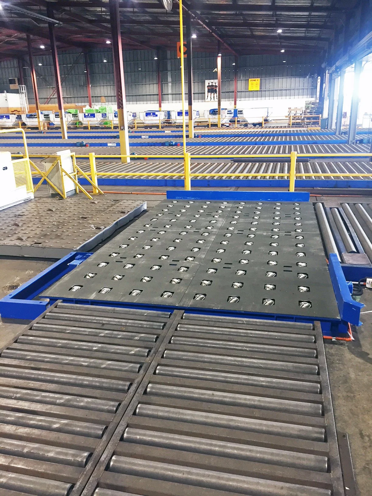 Inside an industrial warehouse with conveyor rollers and a large roller conveyor system in the foreground, yellow safety rails, and a tall ceiling with metal beams. There are signage and equipment in the background.