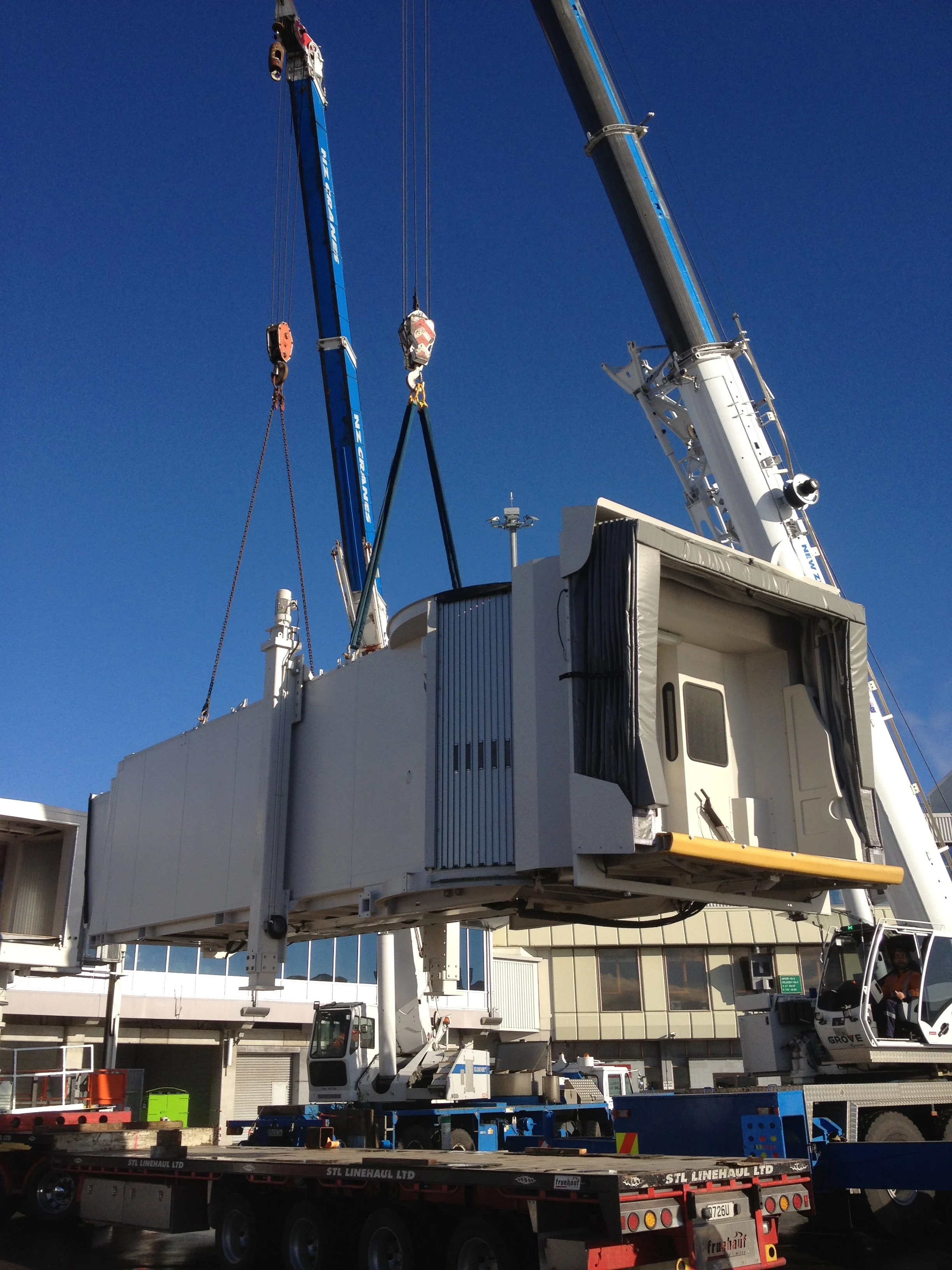 A large crane with a white cab and extendable boom, lifting an airplane passenger boarding bridge against a clear blue sky with a building and other vehicles in the background.