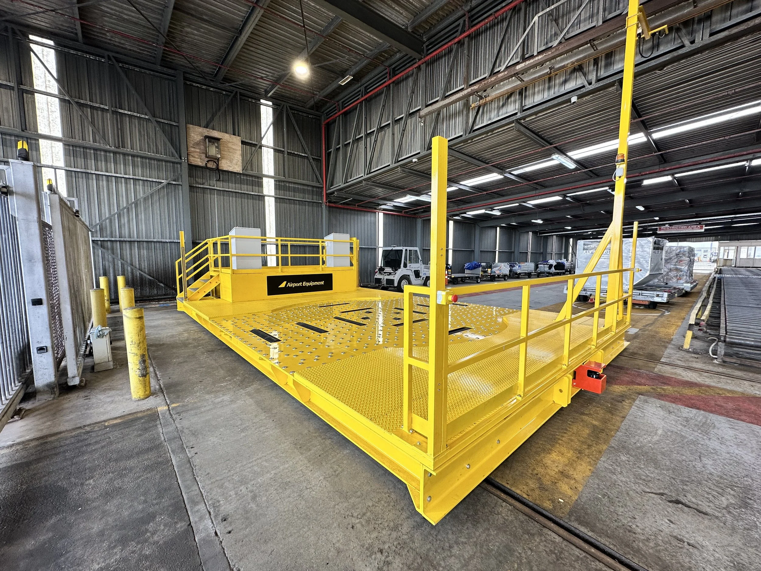 A yellow aircraft service lift inside a large hangar with a metal roof and walls. The lift has a grated platform and safety railings, with some carts and equipment nearby.