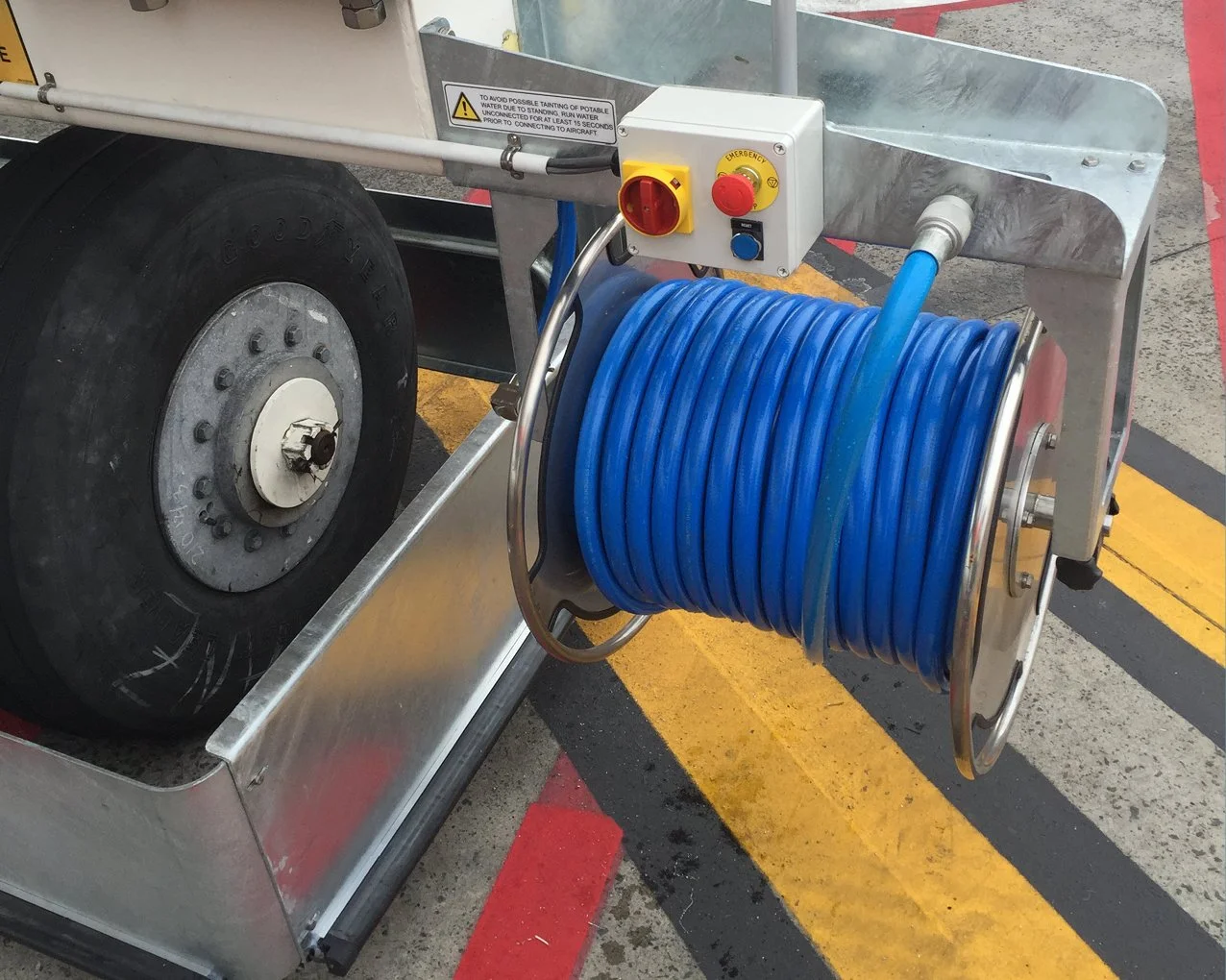 A potable water reel with a blue hose mounted on a metal stand, alongside a control box with emergency stop, power, and control switches, situated on a concrete floor with yellow and black safety lines.