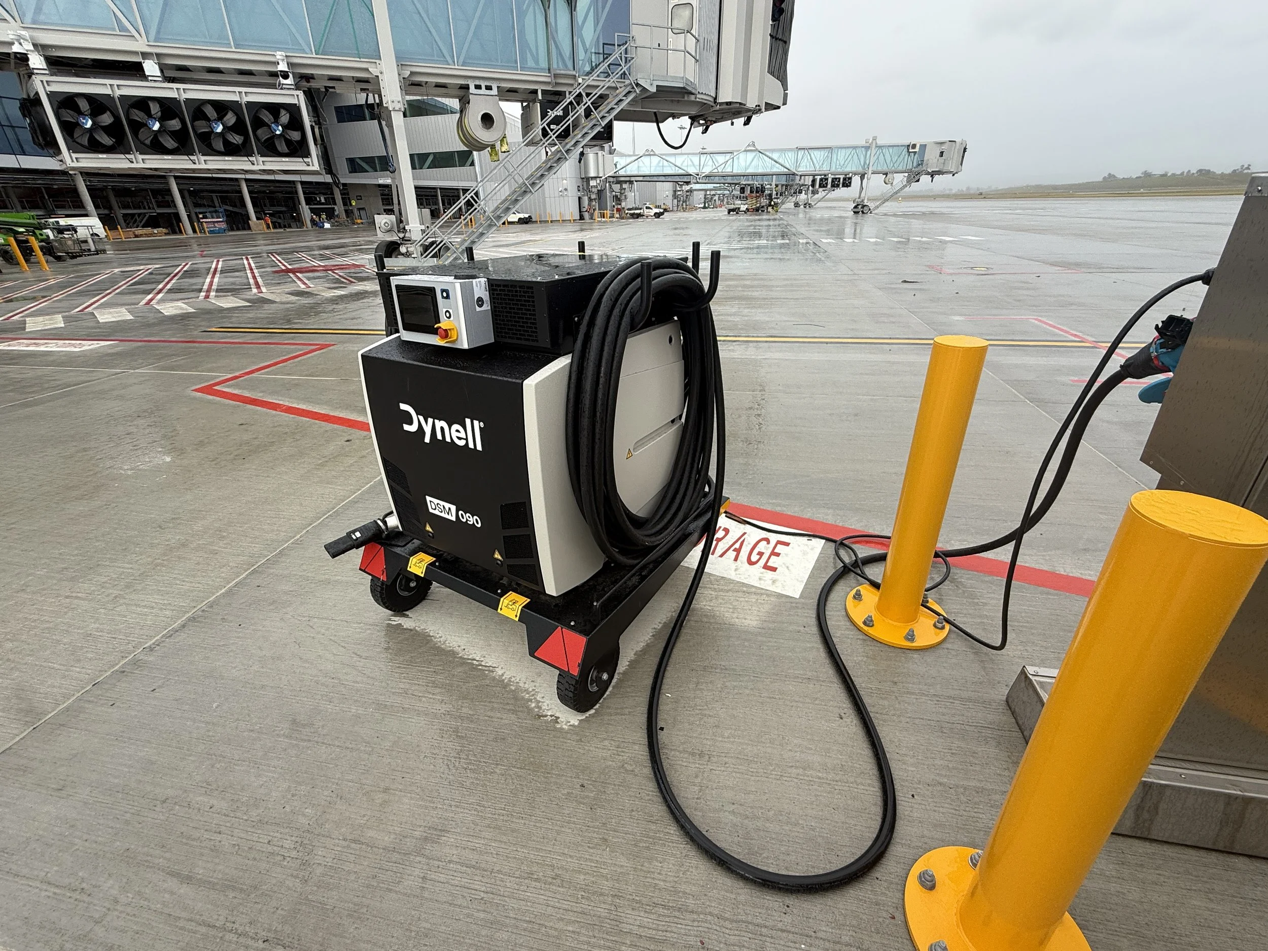 View of an airport tarmac with a ground power unit labeled 'Dynell' connected to an aircraft. In the background, airport jet bridges extend toward the airplane, with some visible airport vehicles and equipment nearby. The scene appears to be rainy or