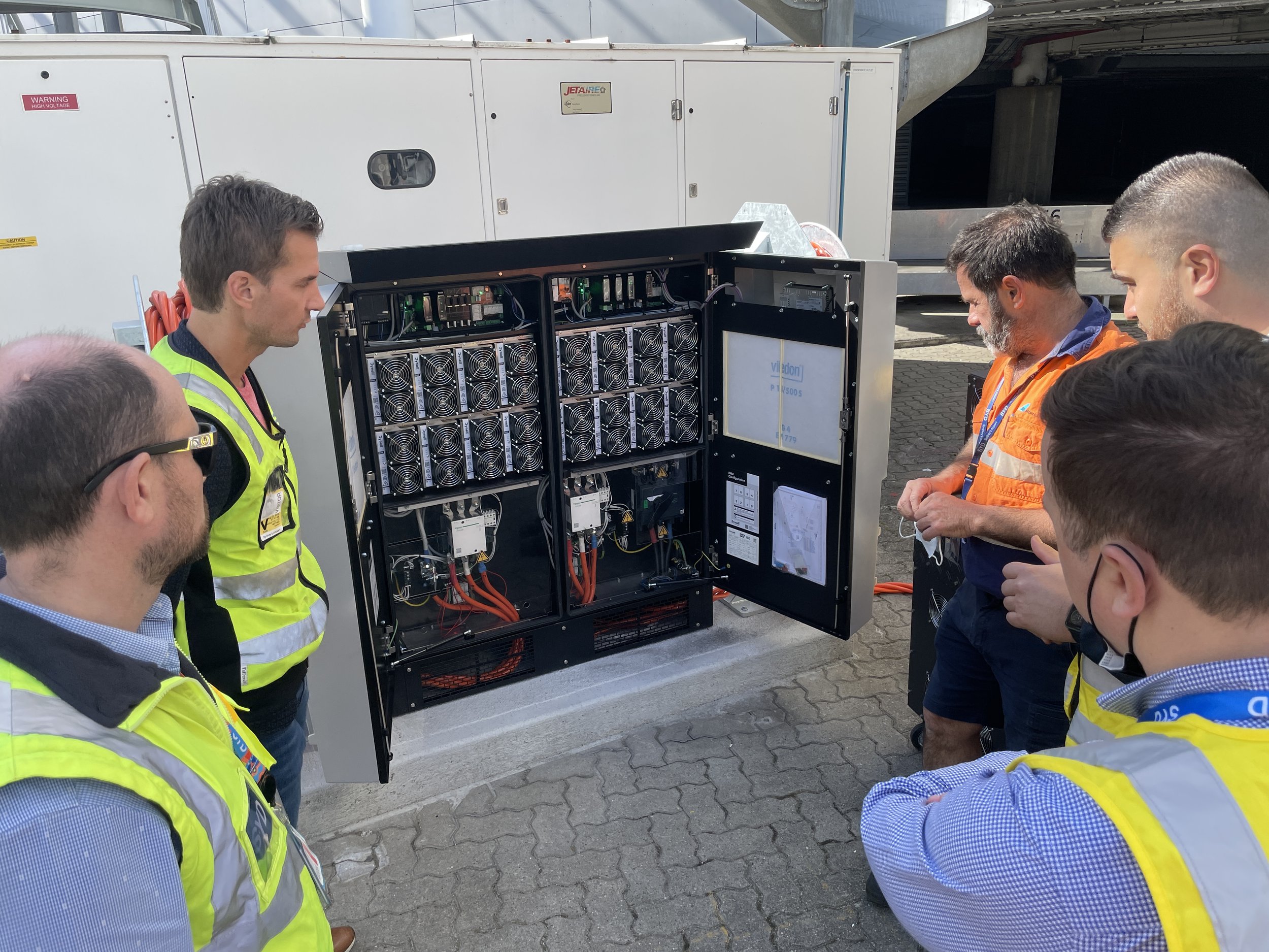 Group of Airport Equipment technicians inspecting an open Dynell System, with cooling fans and electrical components visible, while others observe.