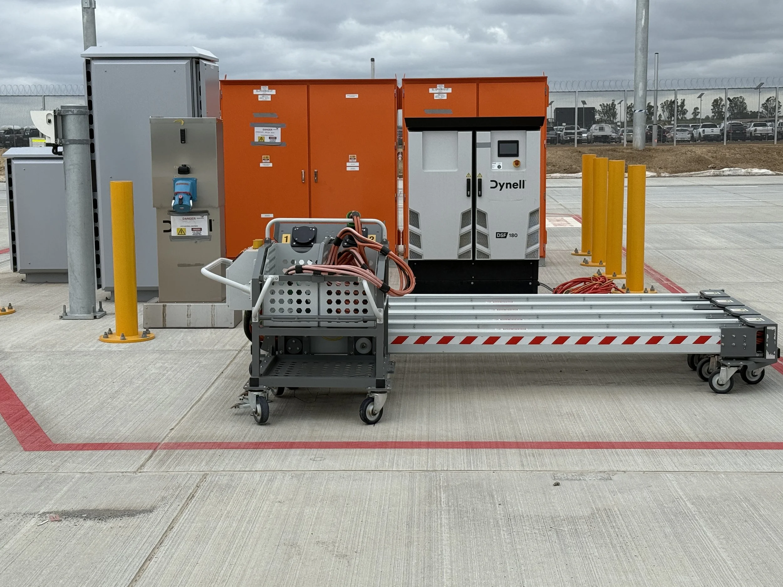 Industrial equipment setup outdoors at an airport, including a Dynell System, electrical cabinets, control panels, orange storage lockers, and yellow safety barriers, with a cloudy sky and parked cars in the background.