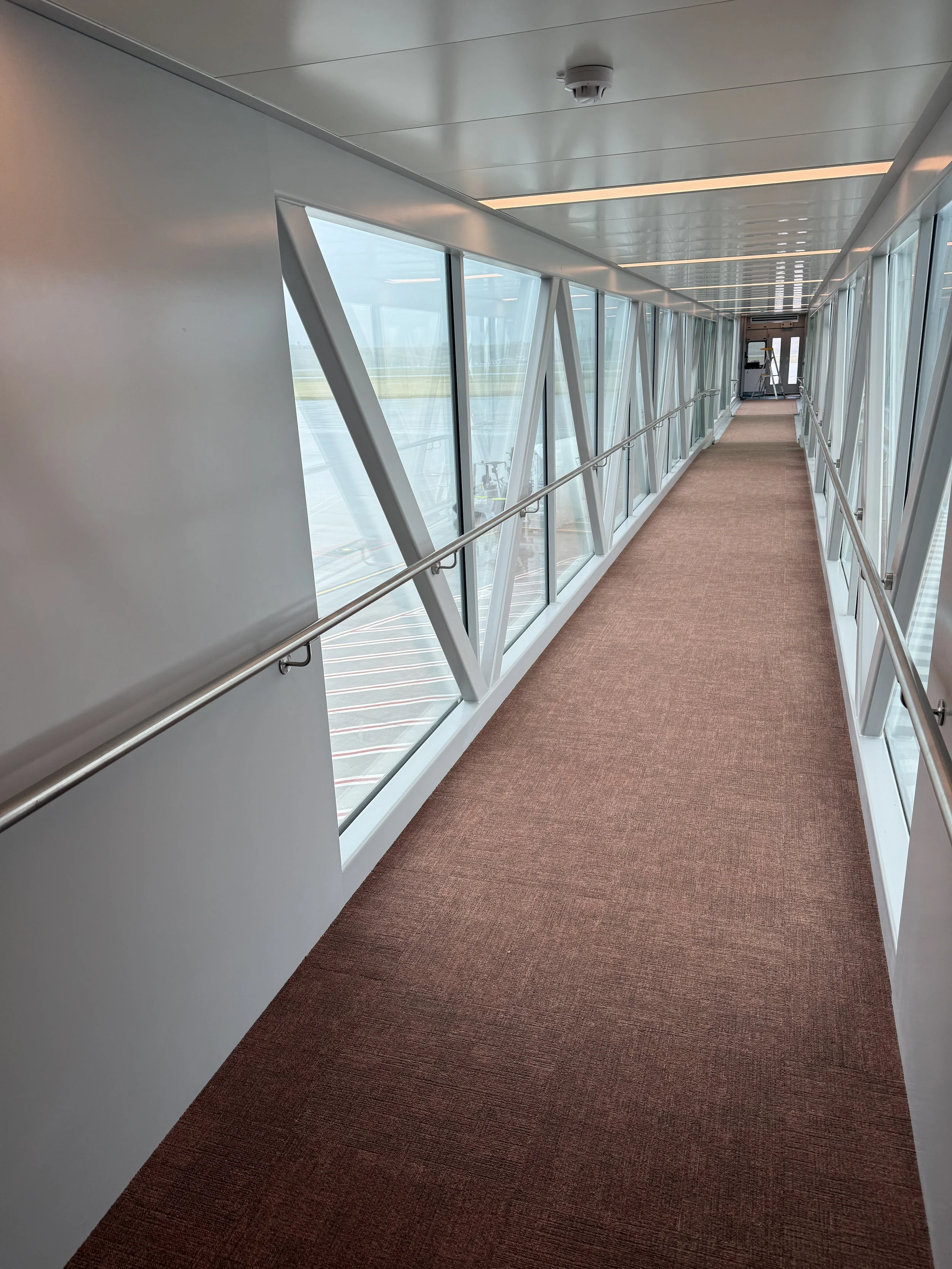 An enclosed passenger boarding bridge with large glass windows on both sides, brown carpeted flooring, and white metal framing, leading to a door at the far end.