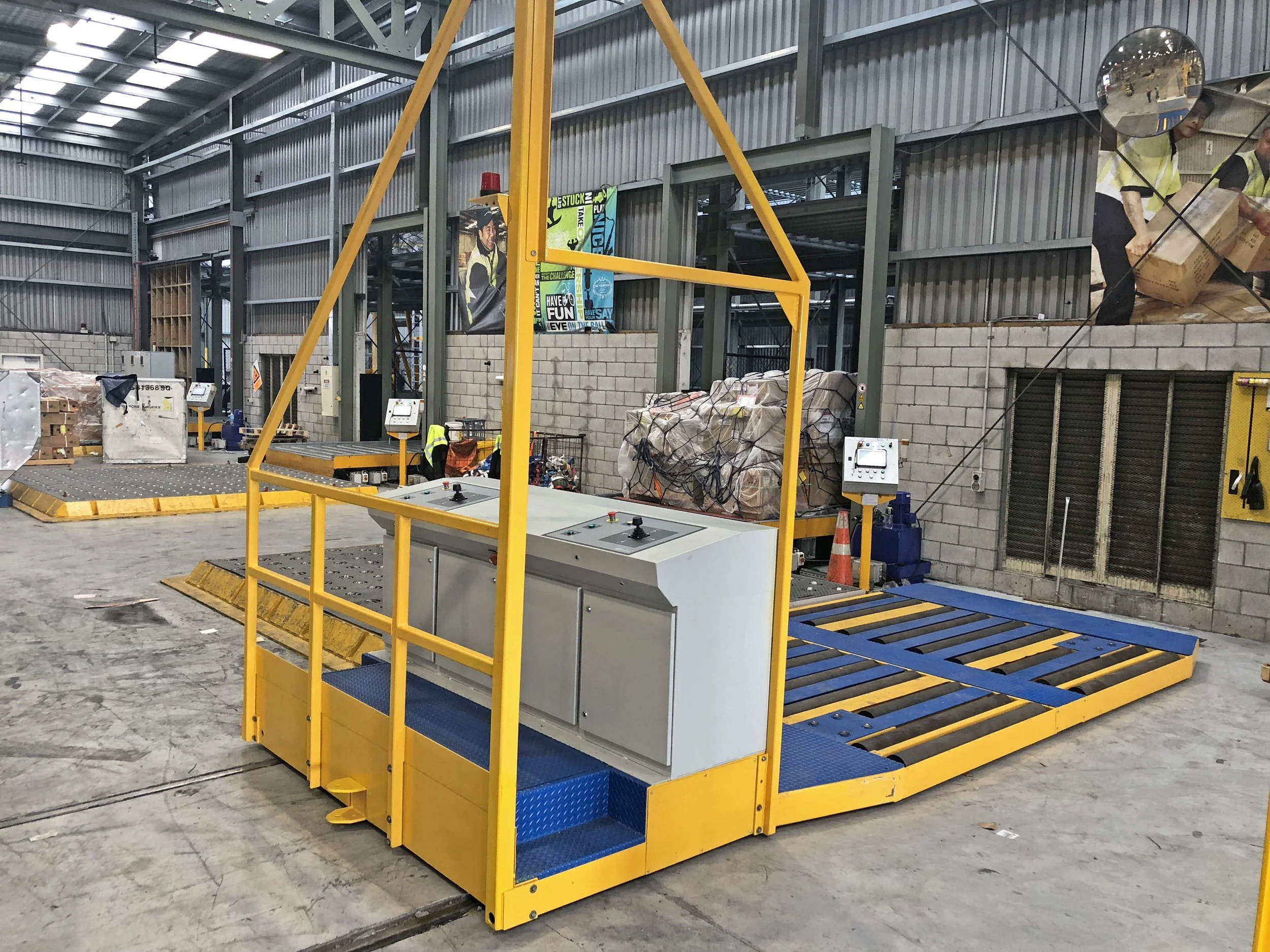 Interior view of a warehouse showing a yellow and blue metal platform with stairs, machinery, stacks of wrapped goods, and workers handling a box.