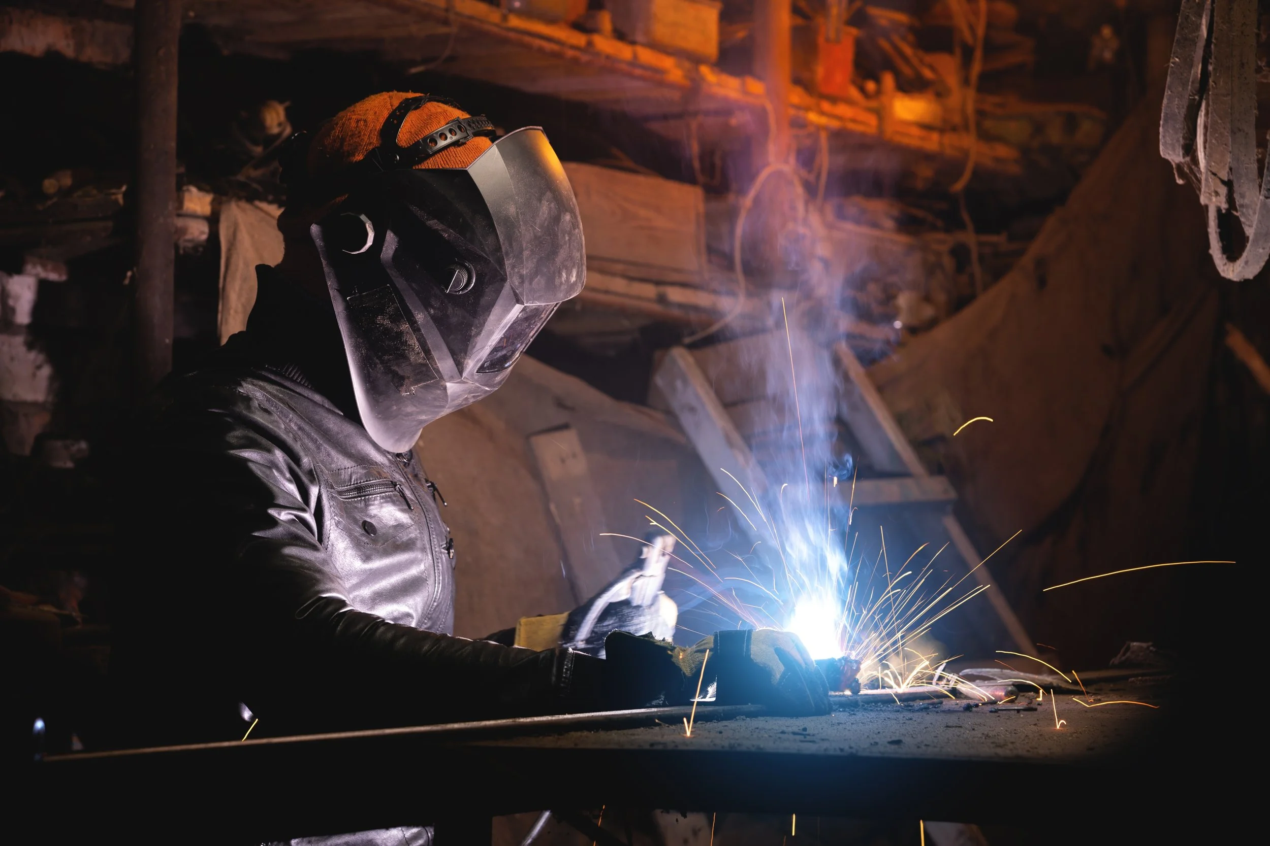 A person welding metal in a workshop, wearing protective gear including a welding helmet, gloves, and a leather jacket, with sparks and bright light from the welding process.