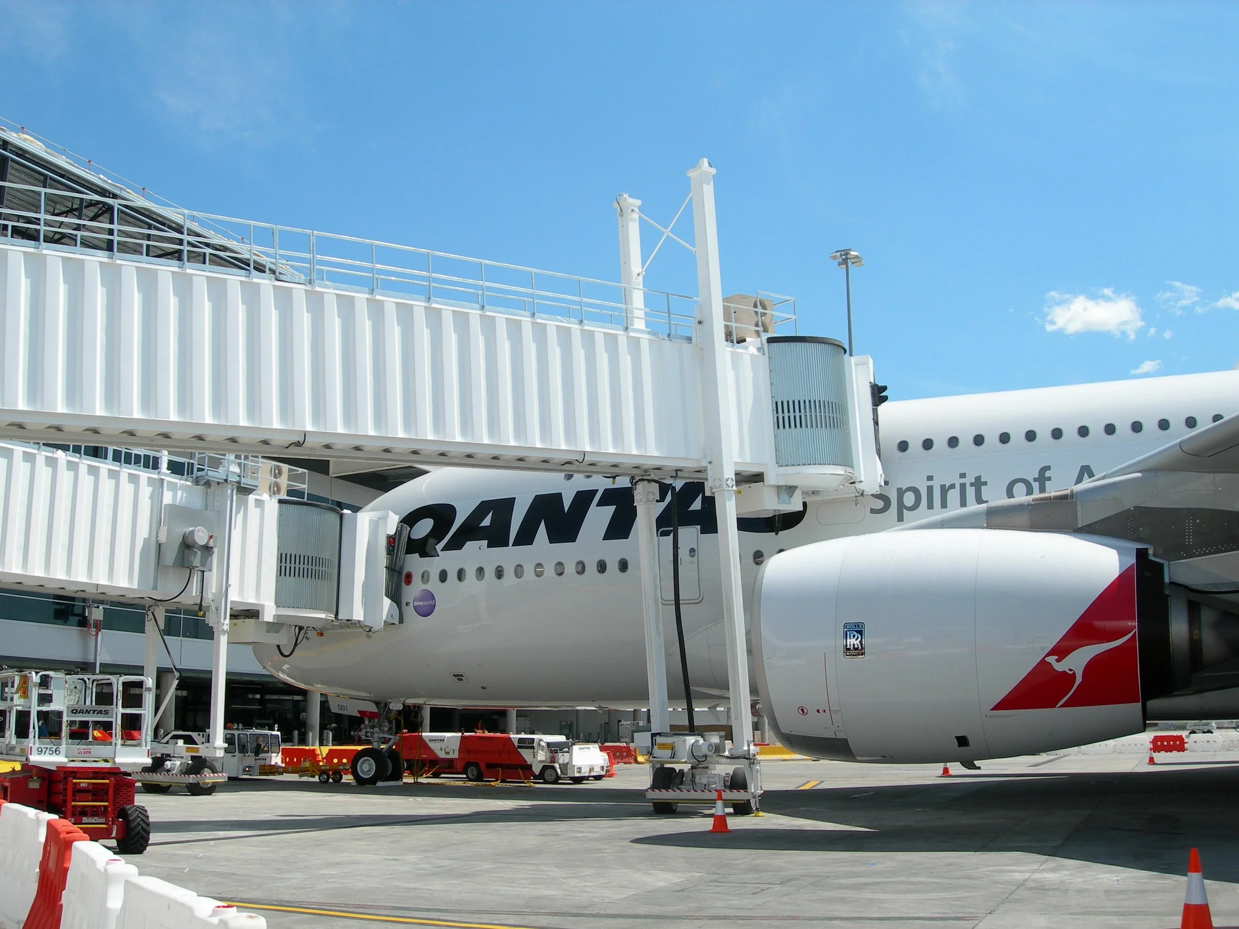 An airplane parked at an airport gate connected to the terminal via a passenger boarding bridge. Ground support equipment and orange safety cones are visible around the aircraft.