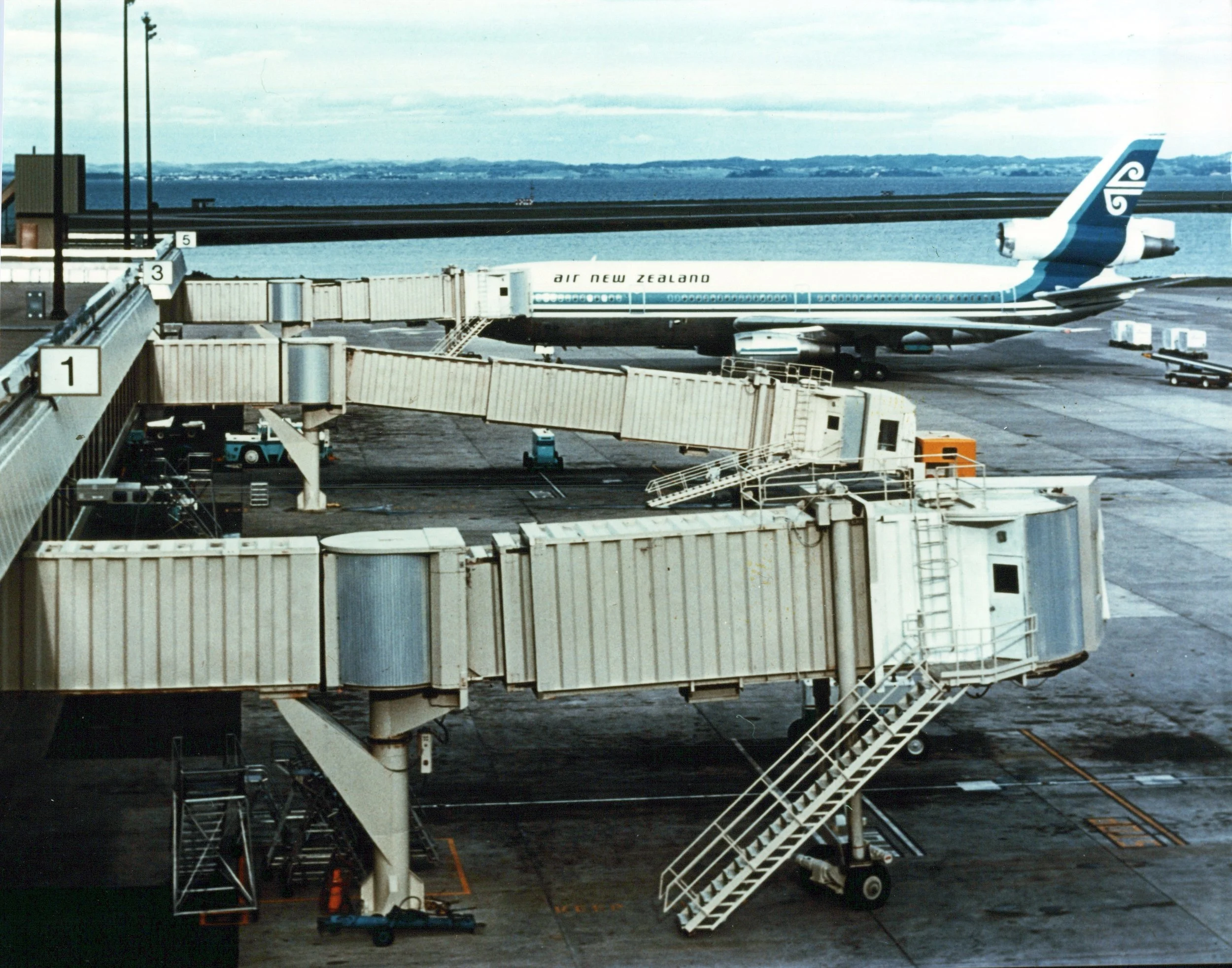A vintage Air New Zealand airplane parked at an airport gate, with boarding bridges, stairs, and airport equipment visible in the foreground. The aircraft is white with blue and green branding, and the background shows a body of water and distant land masses.