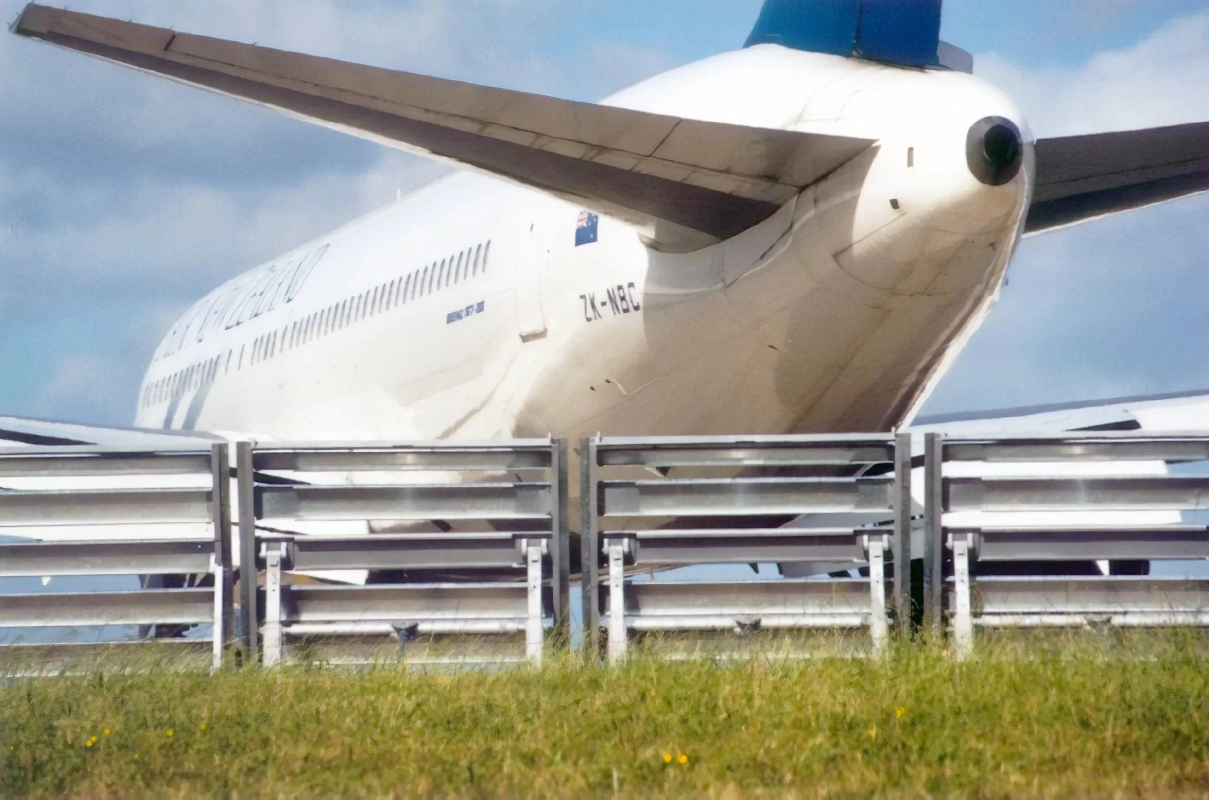 View of the underside of an airplane wing and engine on a runway, with a part of the blast fence and grassy area in the foreground.