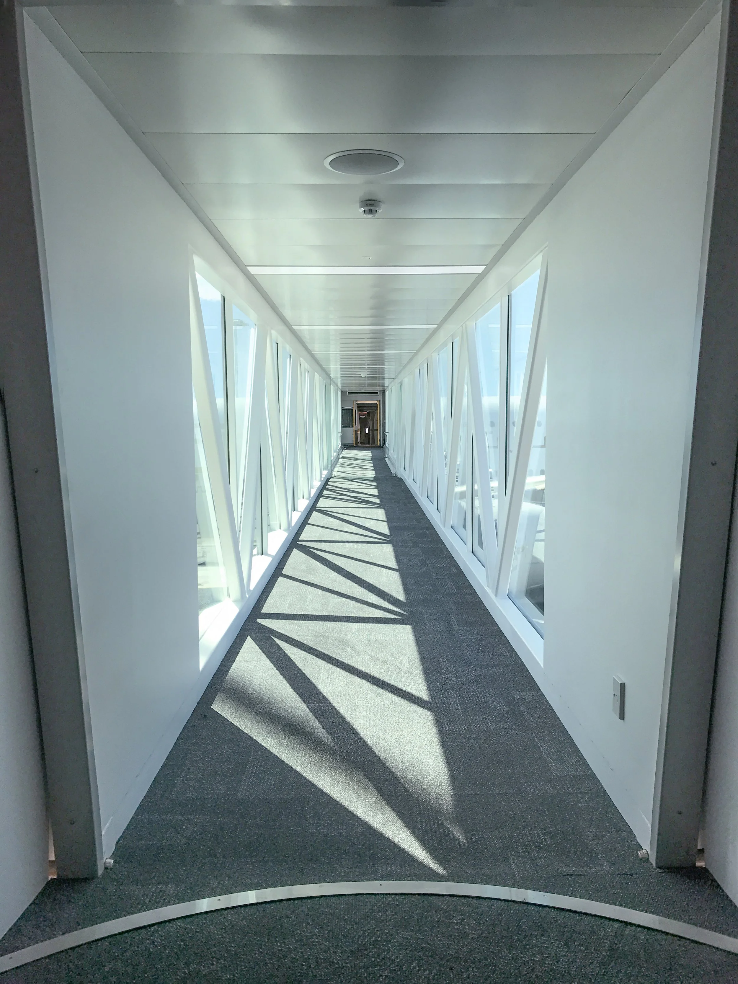 Interior view of a passenger boarding bridge with glass walls on both sides, leading to an exit door, with shadows cast by the window frames on the carpeted floor.