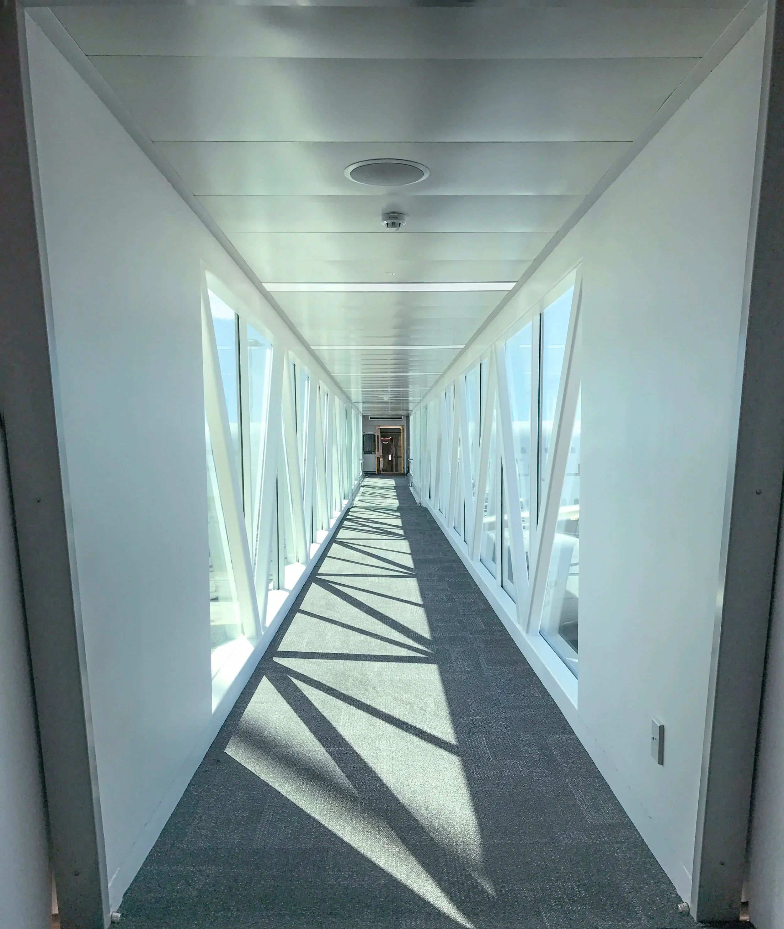 A passenger boarding bridge with large windows on both sides, sunlight casting shadows on the carpeted floor, leading to a doorway in the distance.
