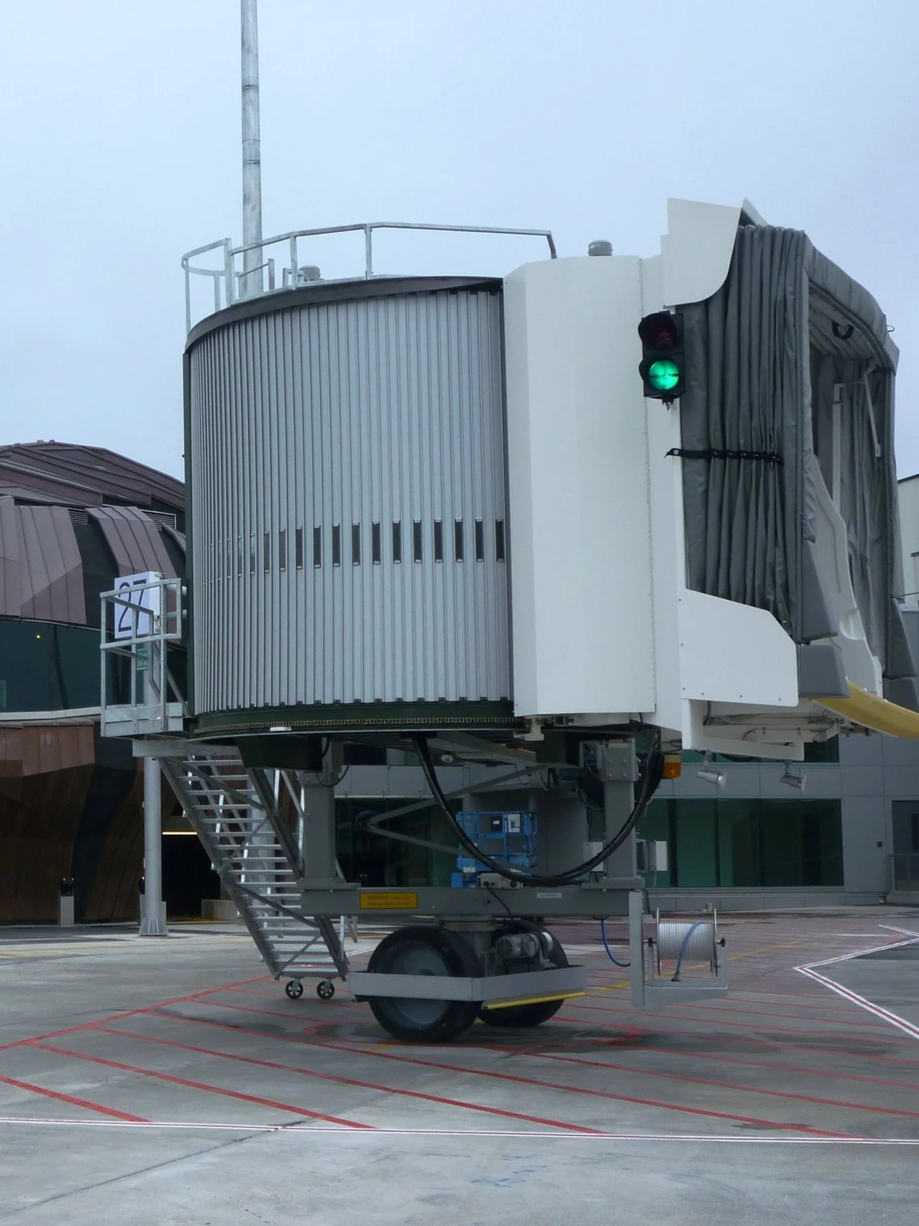 A mobile airport jetbridge with a green traffic light, parked on a tarmac with red and white markings, with buildings in the background.