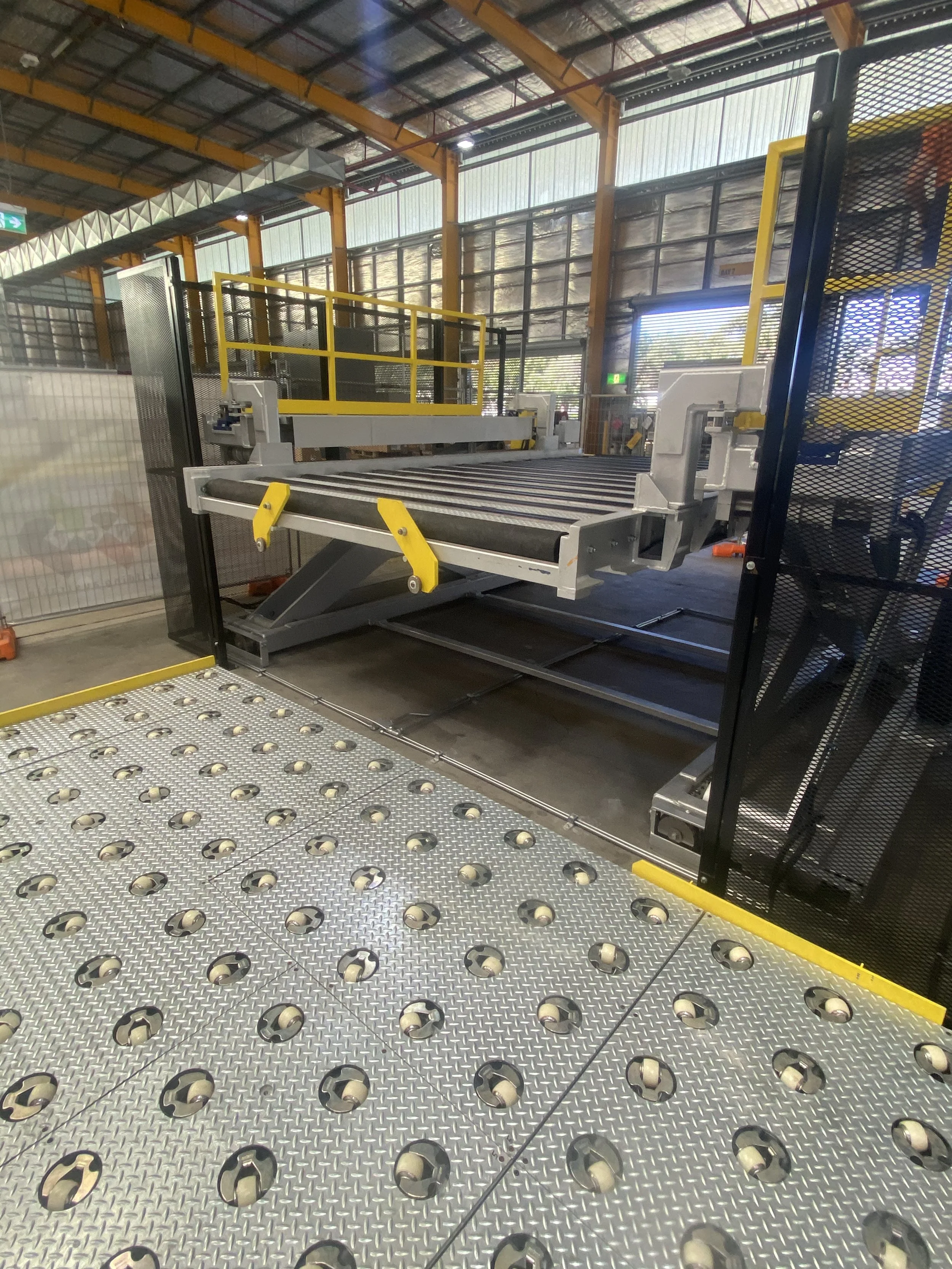 Large industrial machine with rollers and a metal platform, surrounded by safety fencing inside a warehouse or manufacturing facility.