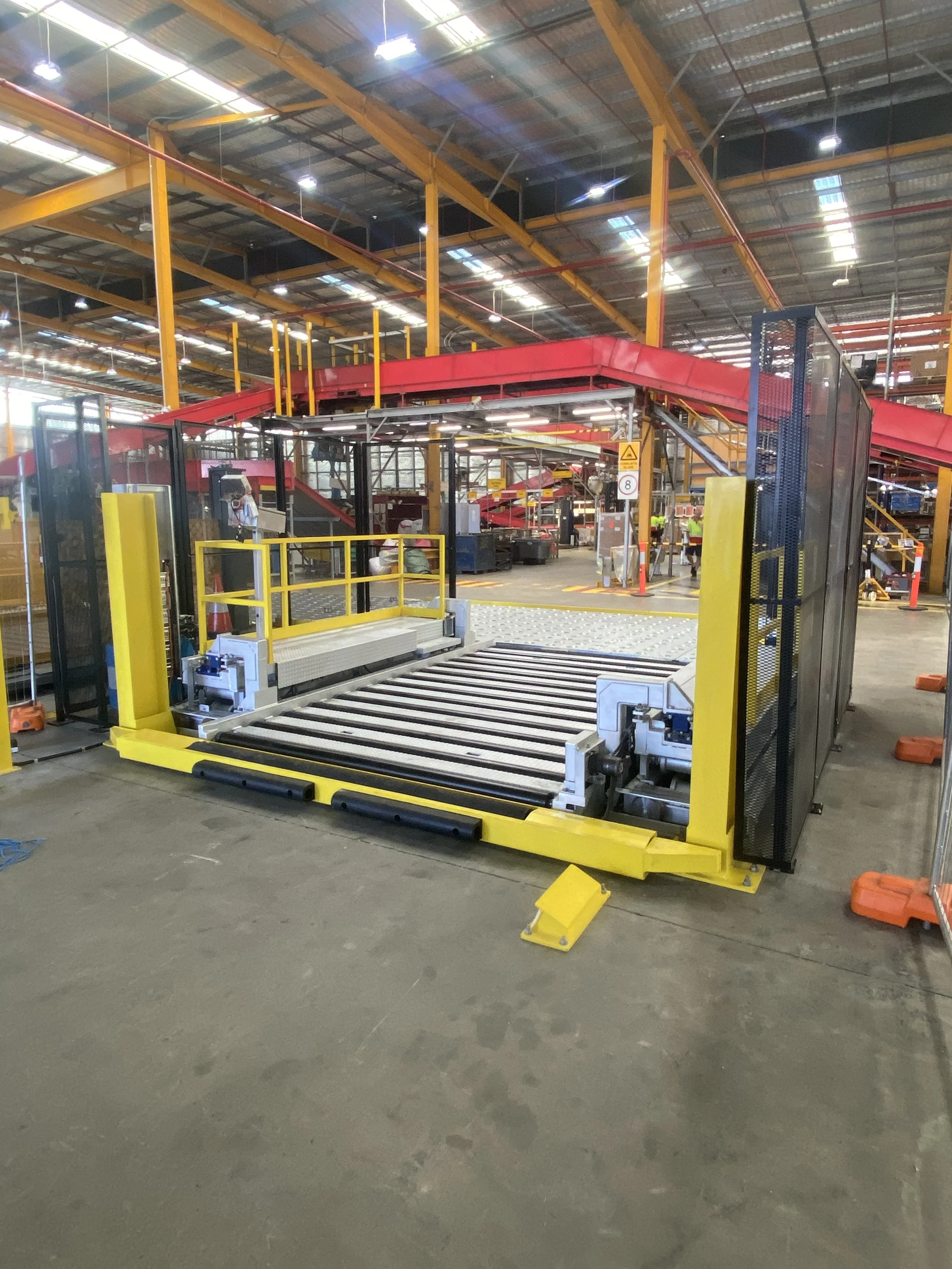 An industrial warehouse with yellow and red machinery used for automated sorting, with conveyor belts and safety barriers, and workers in high-visibility vests in the background.