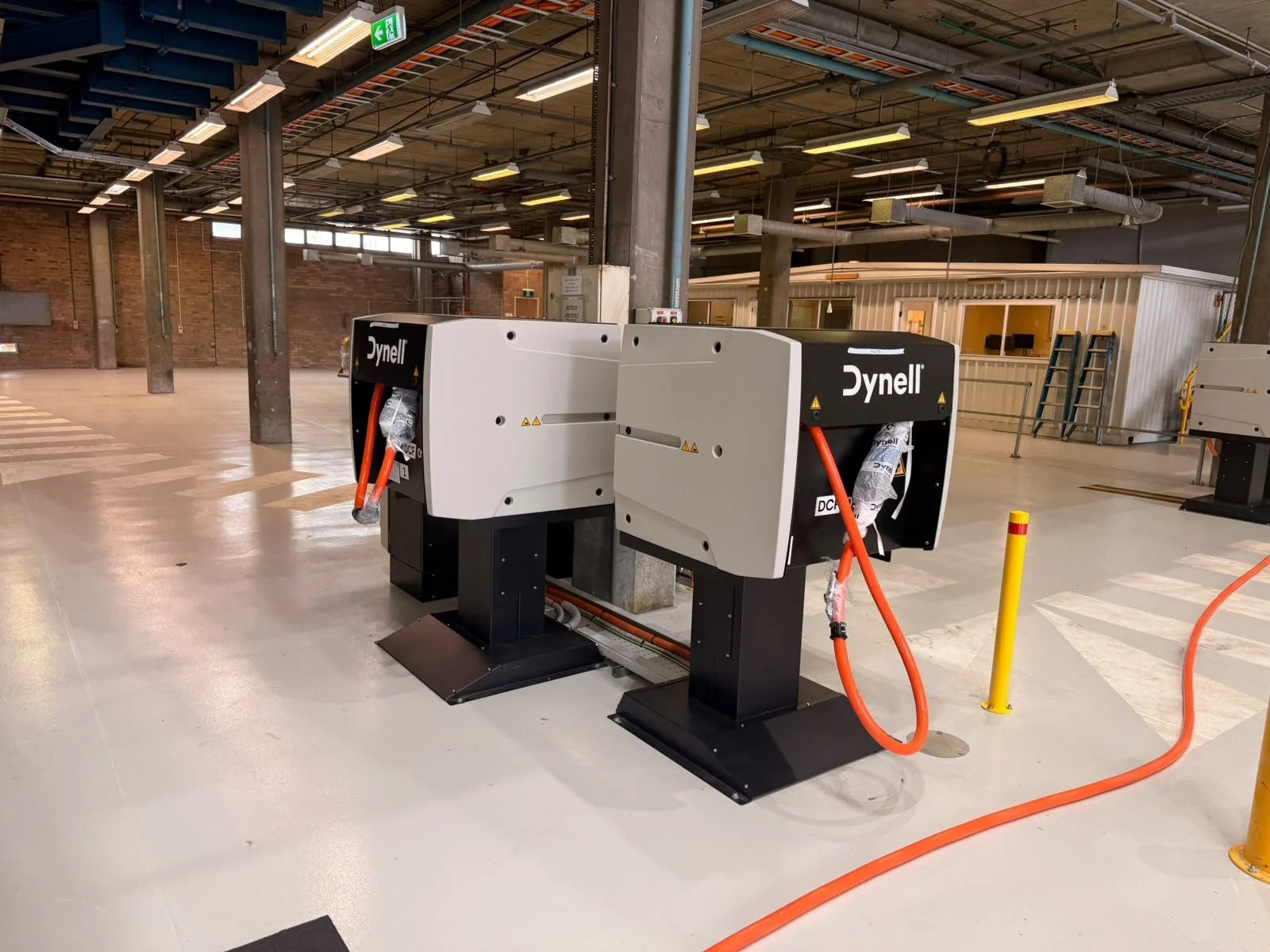 Two Dynell Systems with orange cables in an industrial warehouse setting with high ceilings, exposed pipes, a small office with stairs, and a brick wall in the background.