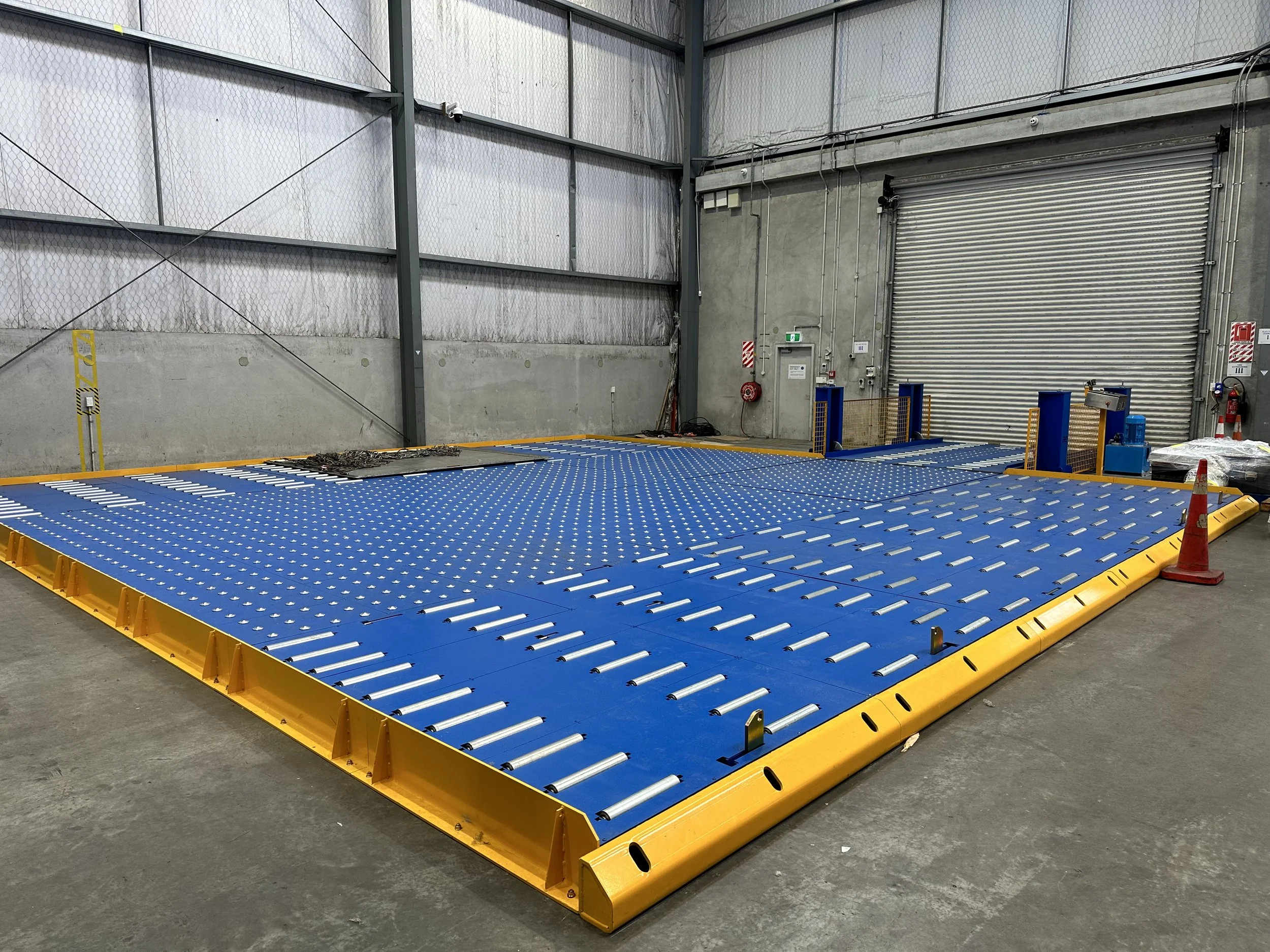 Large industrial blue and yellow manual roller bed in a warehouse, with a large rolling door and concrete wall in the background.