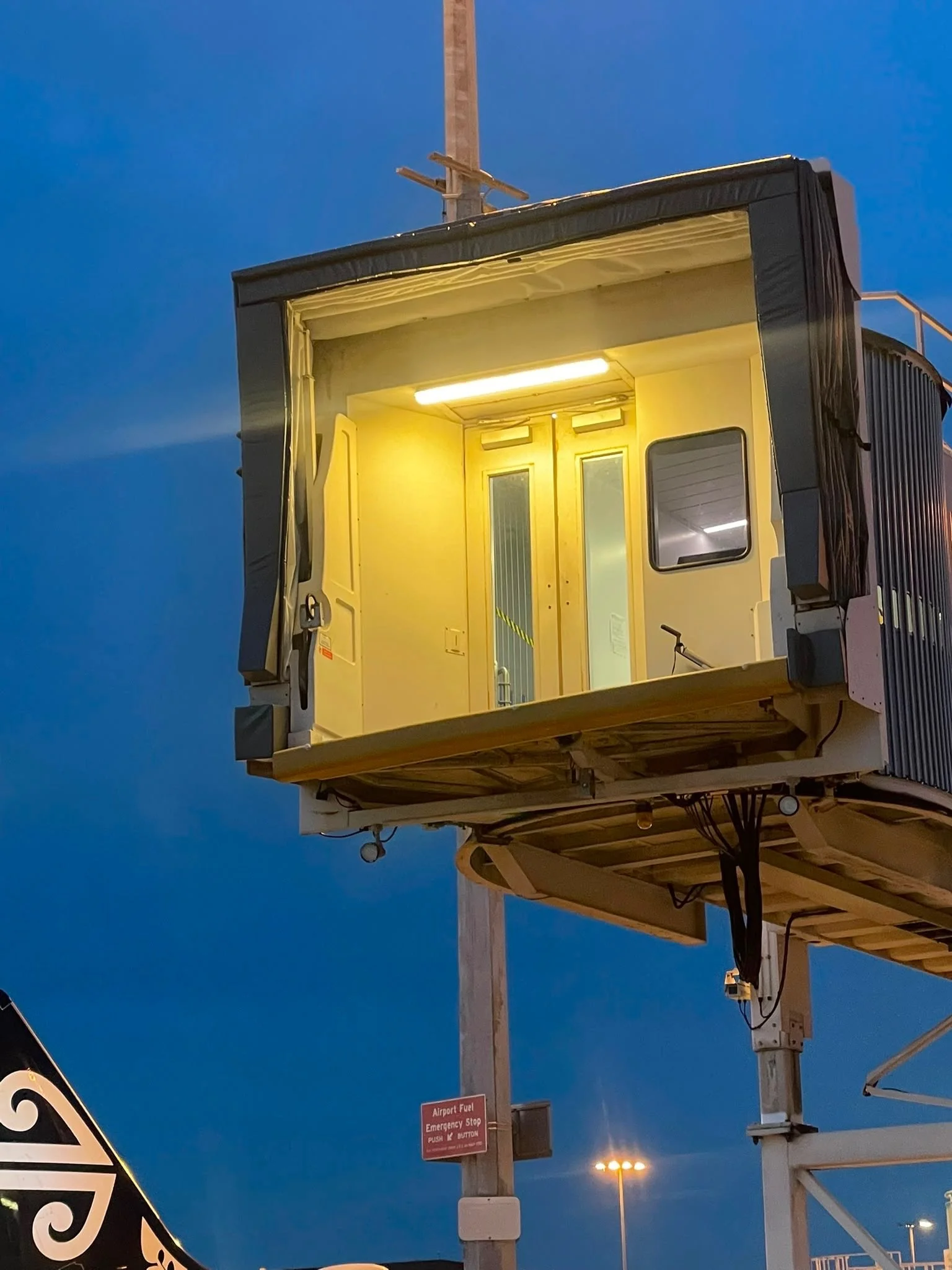 Airport passenger boarding bridge with yellow and black lighting, not yet connected to an aircraft. The scene is set during early evening or dusk with a darkening sky.