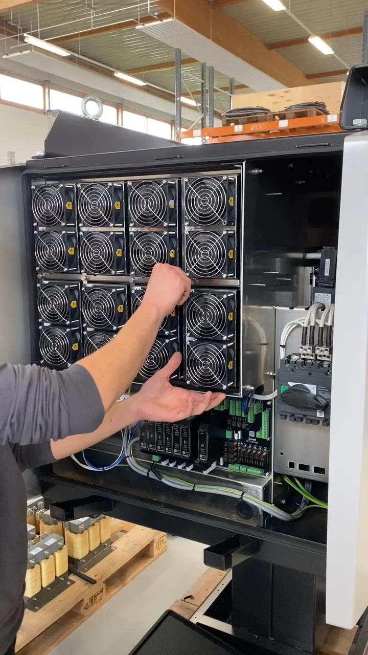 A technician installing or maintaining cooling fans inside a Dynell System. The cabinet contains multiple metal fans with protective grill covers and various wiring and electronic components.