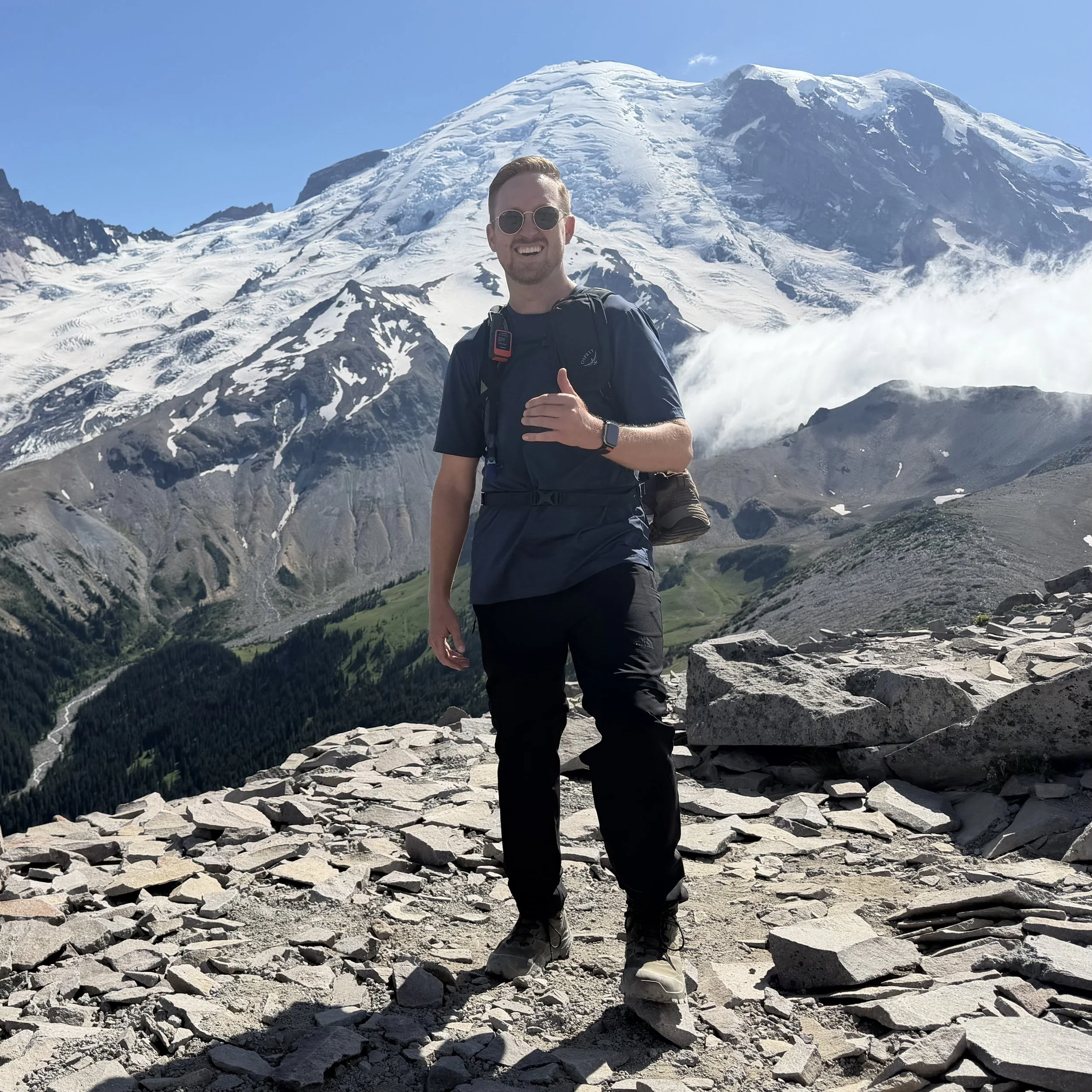 Schro hiking across rocky terrain with a snow covered mountain,  Mount Rainier, in the distant background