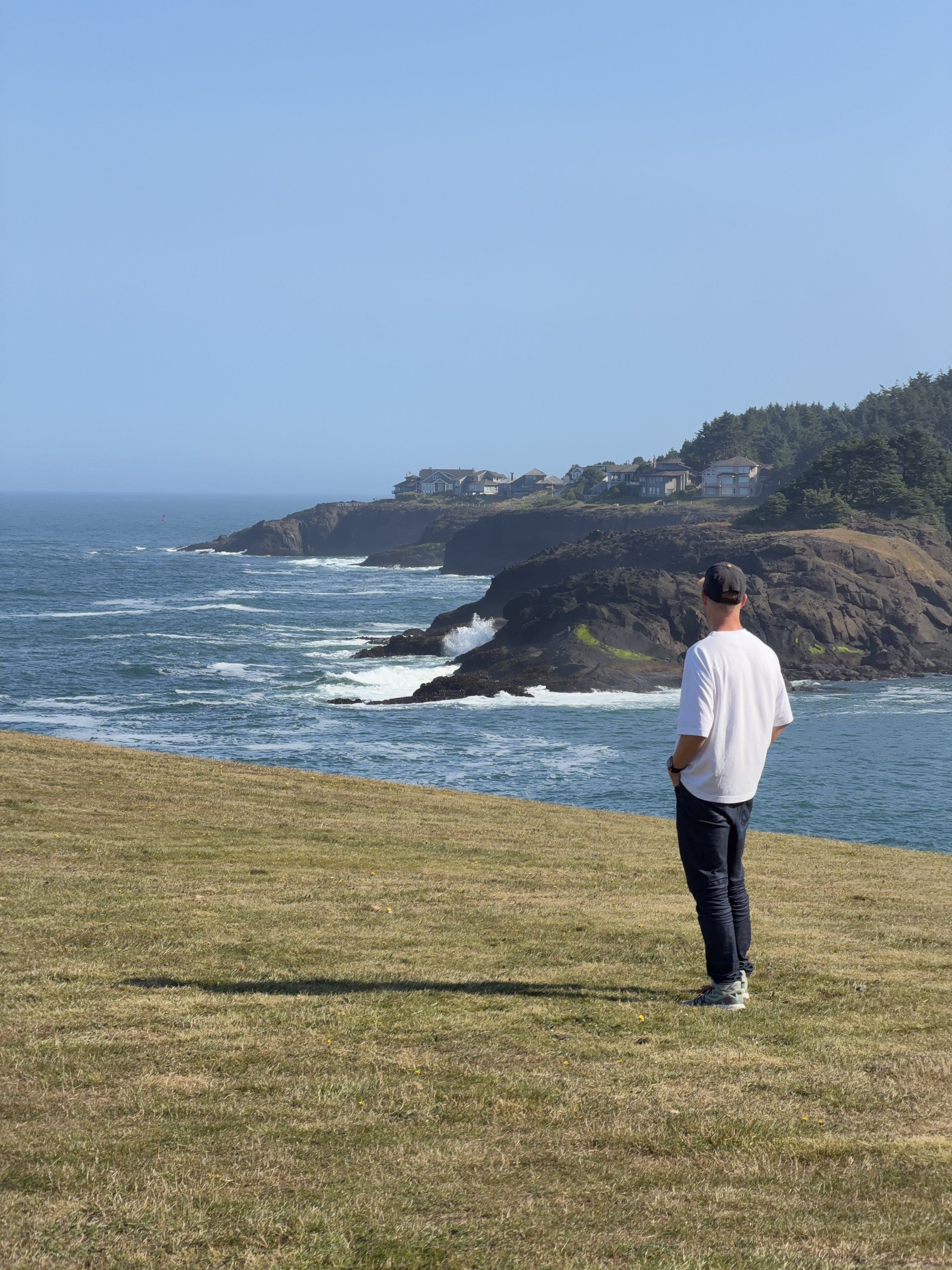 Schro is wearing a white shirt, blue jeans, and a baseball cap stands on a grassy area along the pacific coast in Oregon, overlooking the ocean with rocks and houses on the hillside in the distance under a clear blue sky.