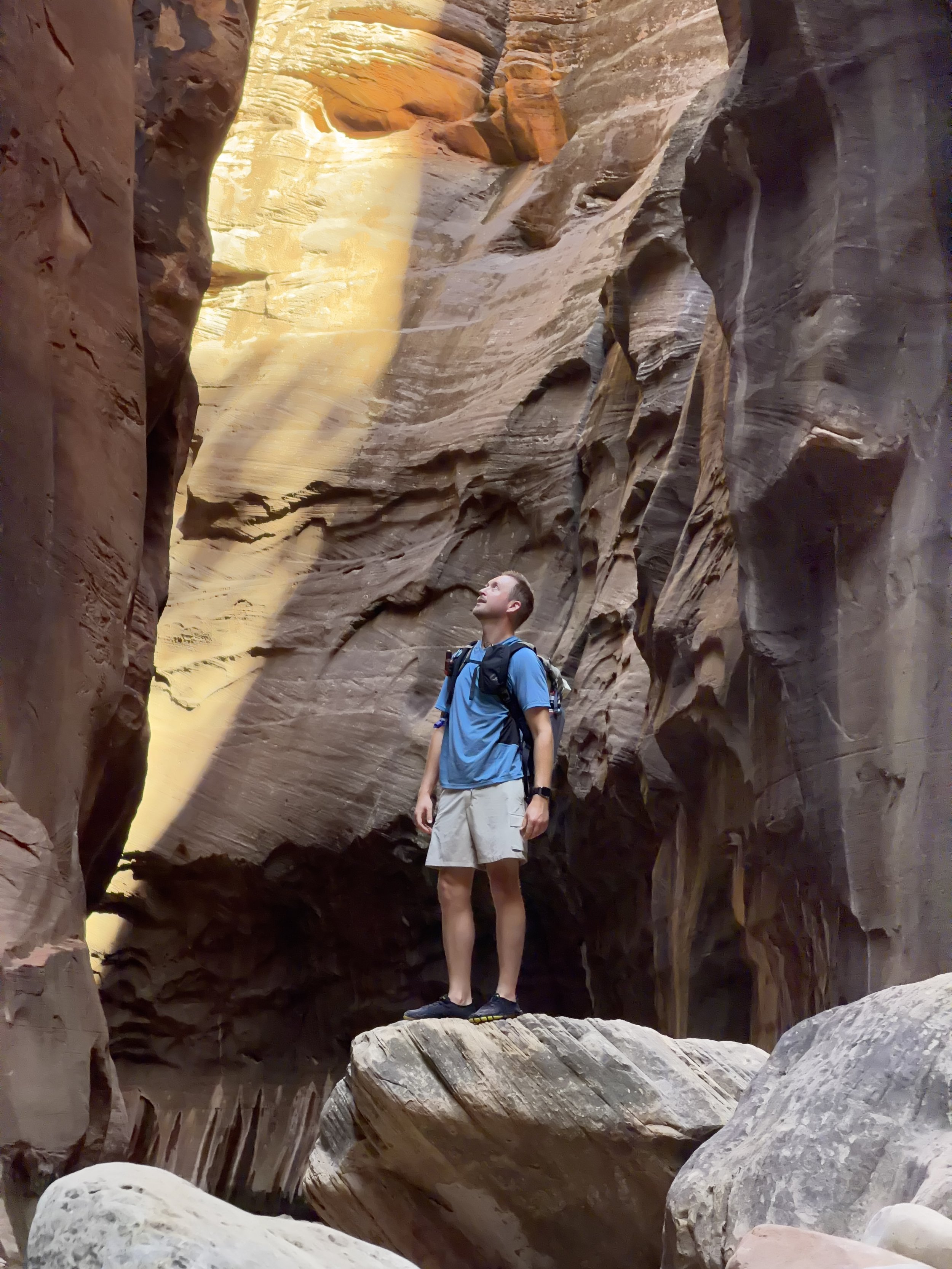 Schro in a blue shirt and beige shorts with a backpack is standing in the Narrows canyon in Zion National Park. He is looking up at the canyon walls, and there is a beam of sunlight illuminating part of the rock face.