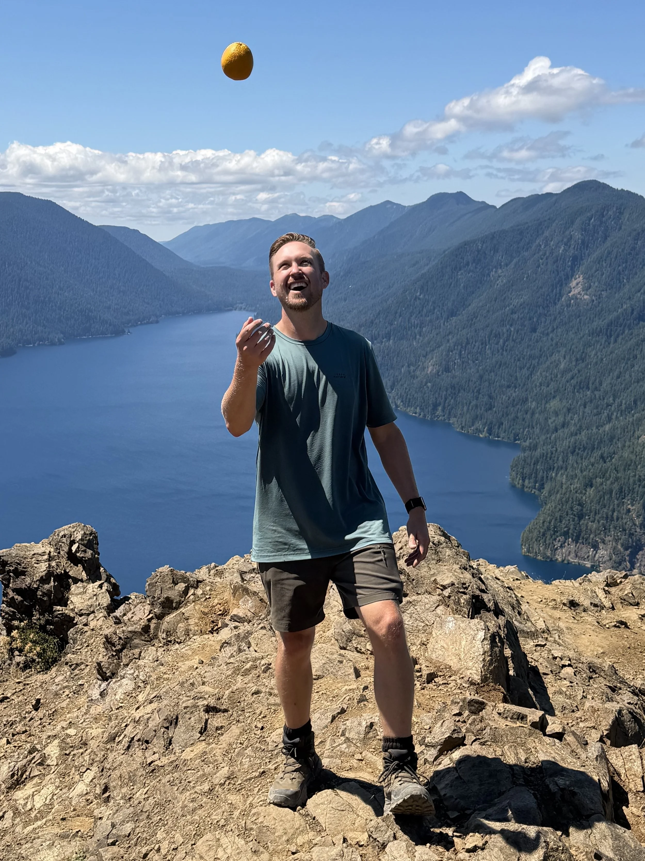 Schro playfully tossing an orange in the air from a mountain above the blue alpine lake, Crescent Lake in Olympic National Park