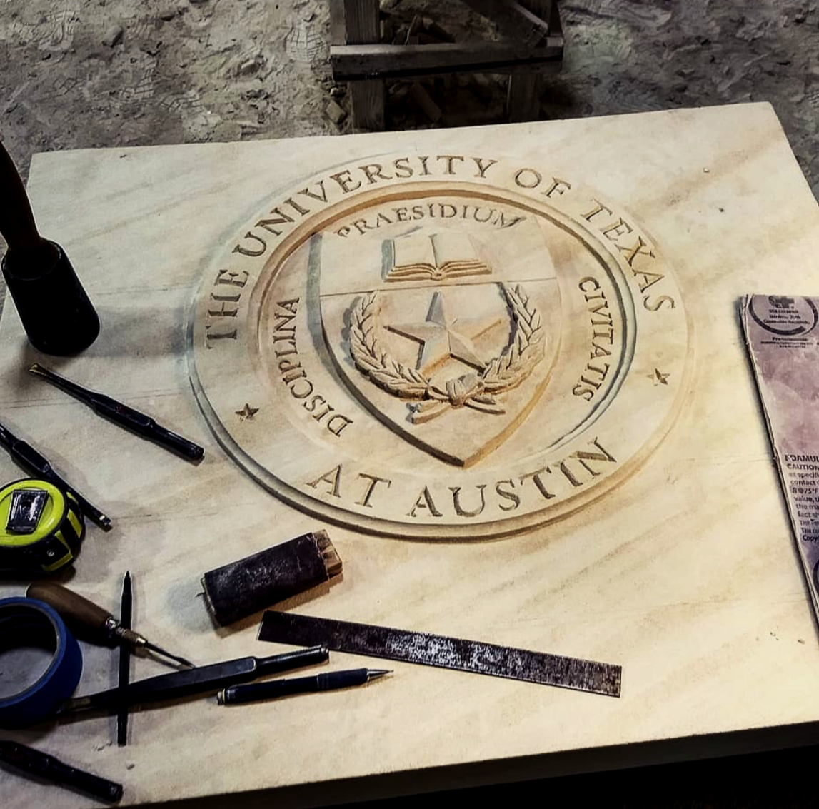 Wooden plaque with the emblem of the University of Texas at Austin being engraved, surrounded by tools and materials for carving.