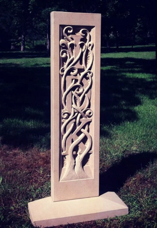 A carved stone sculpture of the letters "IDK" intertwined trees and vines standing outdoors 