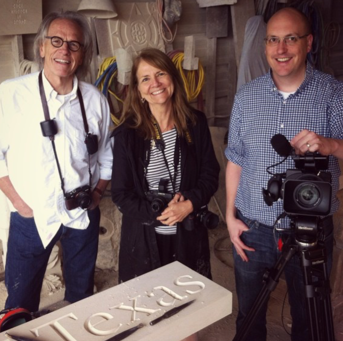 Three smiling people with cameras around their necks standing behind a table with the word 'VOTCAS' and a pen on it, inside a room with wooden walls, wires, and equipment in the background.