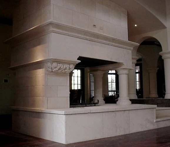 A large limestone fireplace with intricate architectural details, supported by columns, inside a spacious room with arched doorways and tall windows.