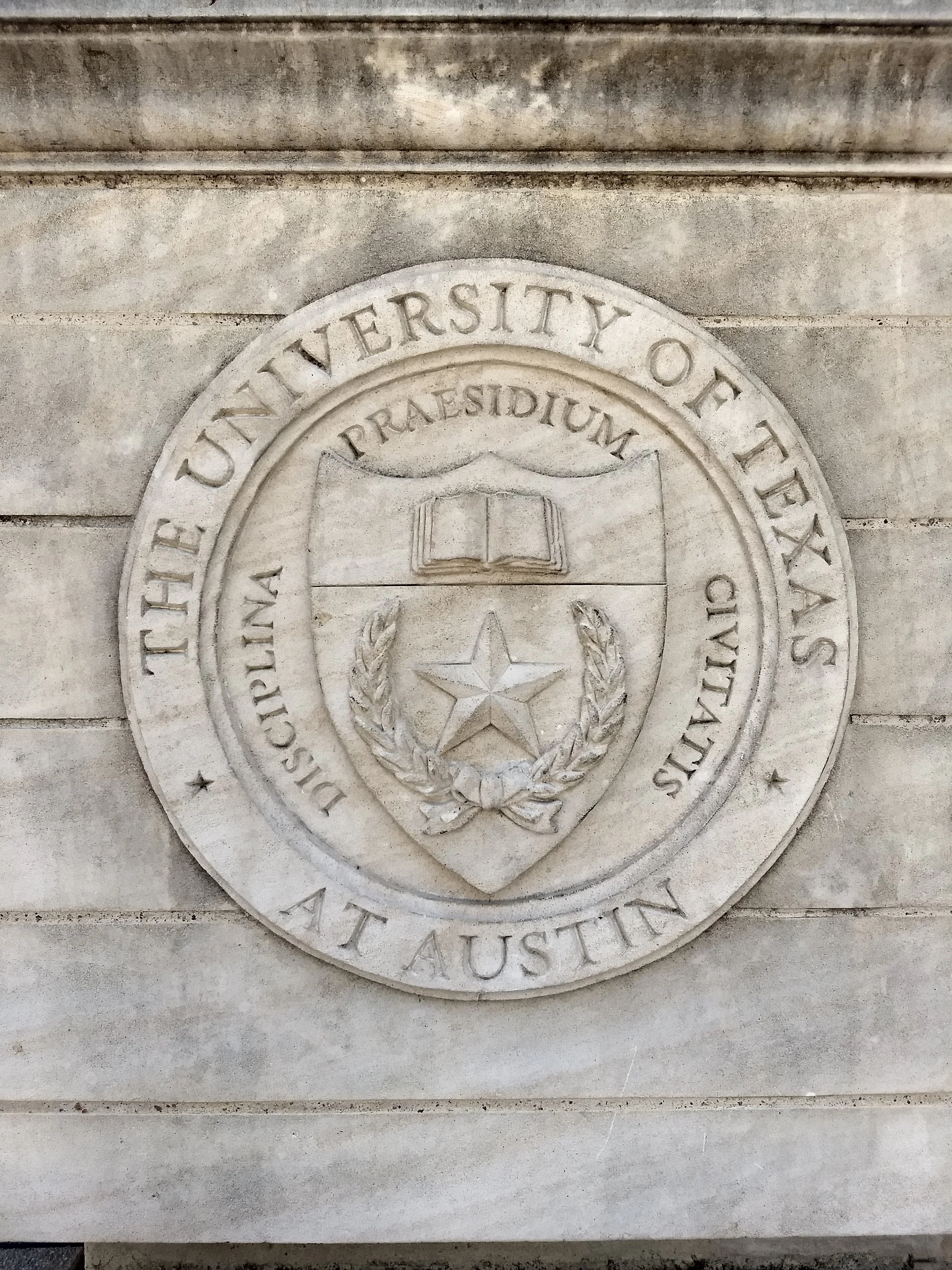 Close-up of a carved seal on a stone wall with the words, 'THE UNIVERSITY OF TEXAS AT AUSTIN,' surrounding a shield with an open book, a star, and a wreath.