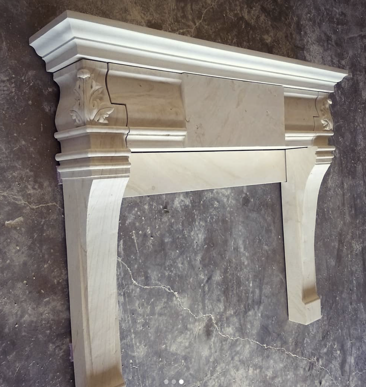 A close-up of a cream-colored marble fireplace mantel with ornate carved details, resting on a cracked concrete floor.