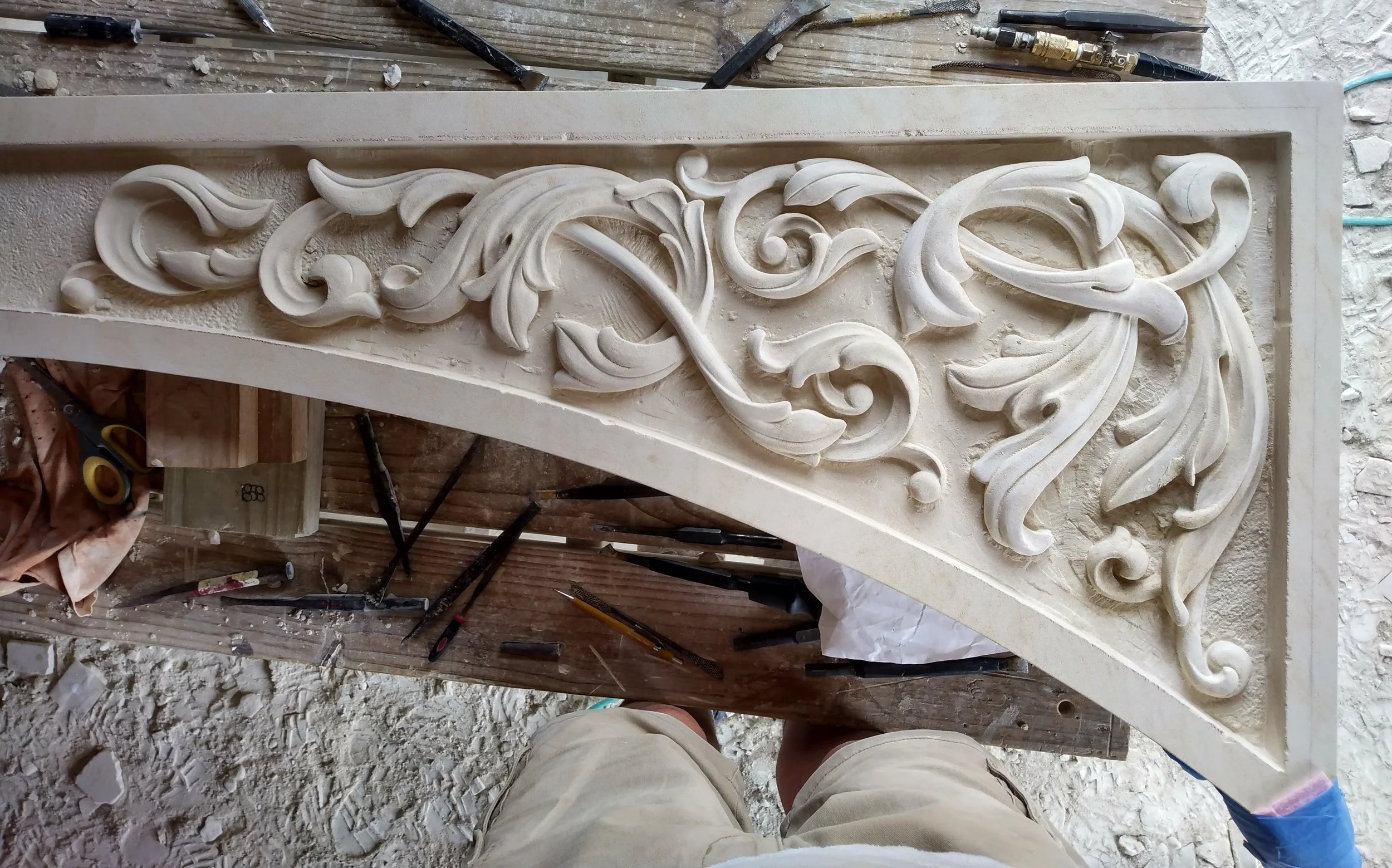 Close-up view of an intricate, ornate carving with floral and scrollwork patterns in a light-colored stone,  with tools and dust around, suggesting ongoing sculpting work.