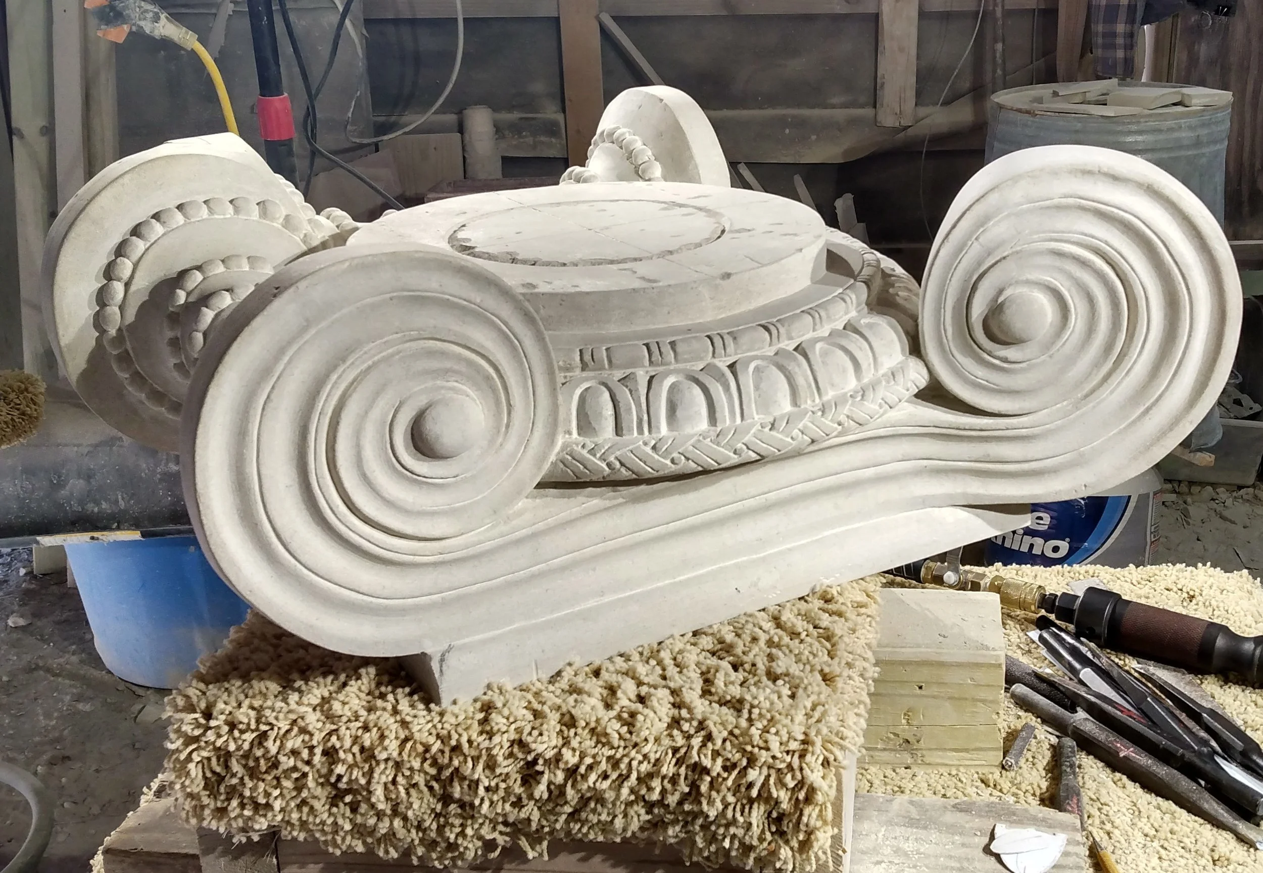 A classical carved stone ionic capital in progress, featuring intricate scroll and bead designs, surrounded by stone carving tools and dust at Bartlett Stone Carving workshop