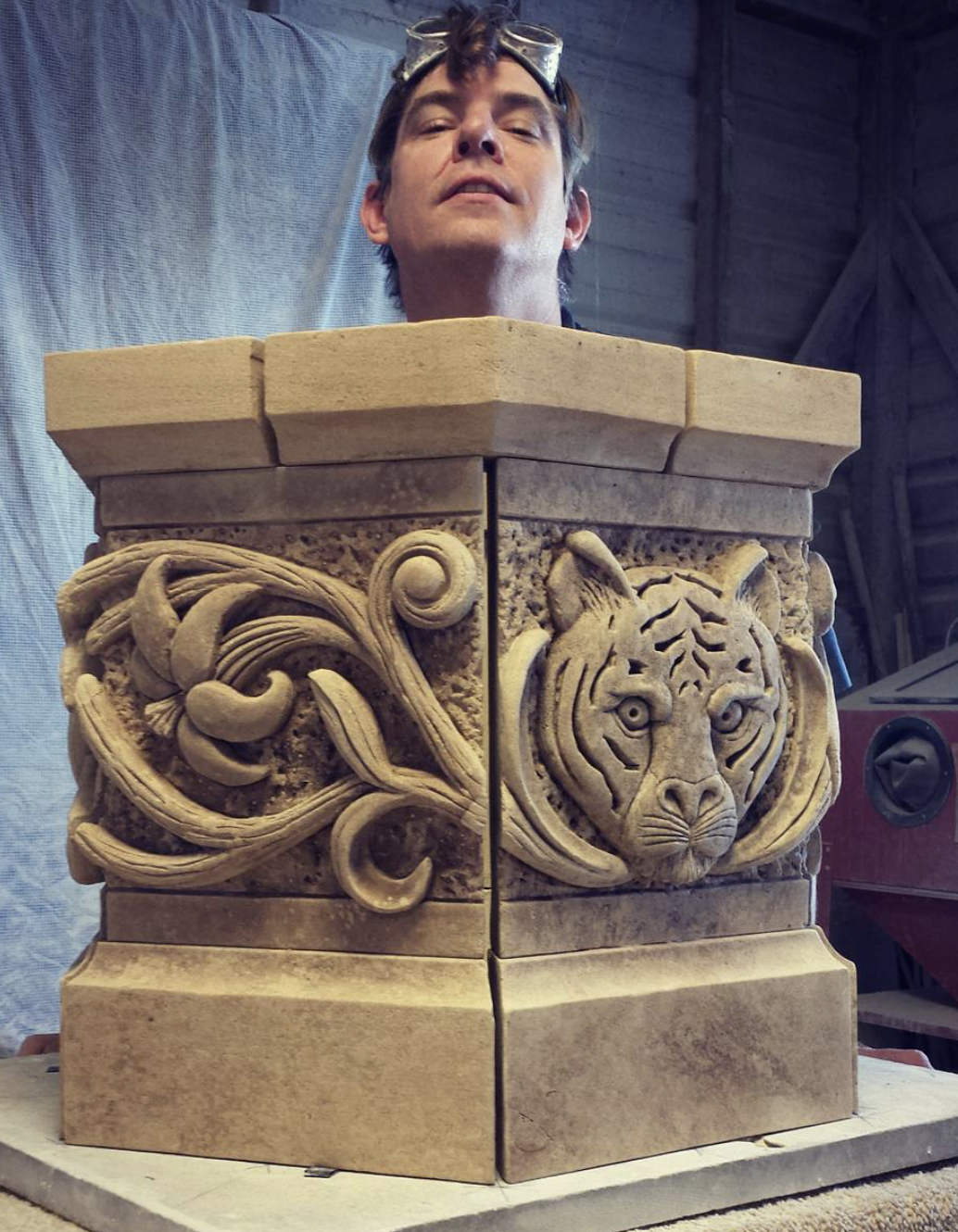 A man wearing safety goggles is seen from below, standing behind intricately carved stone blocks. The carvings feature a tiger's face and ornate swirling patterns.