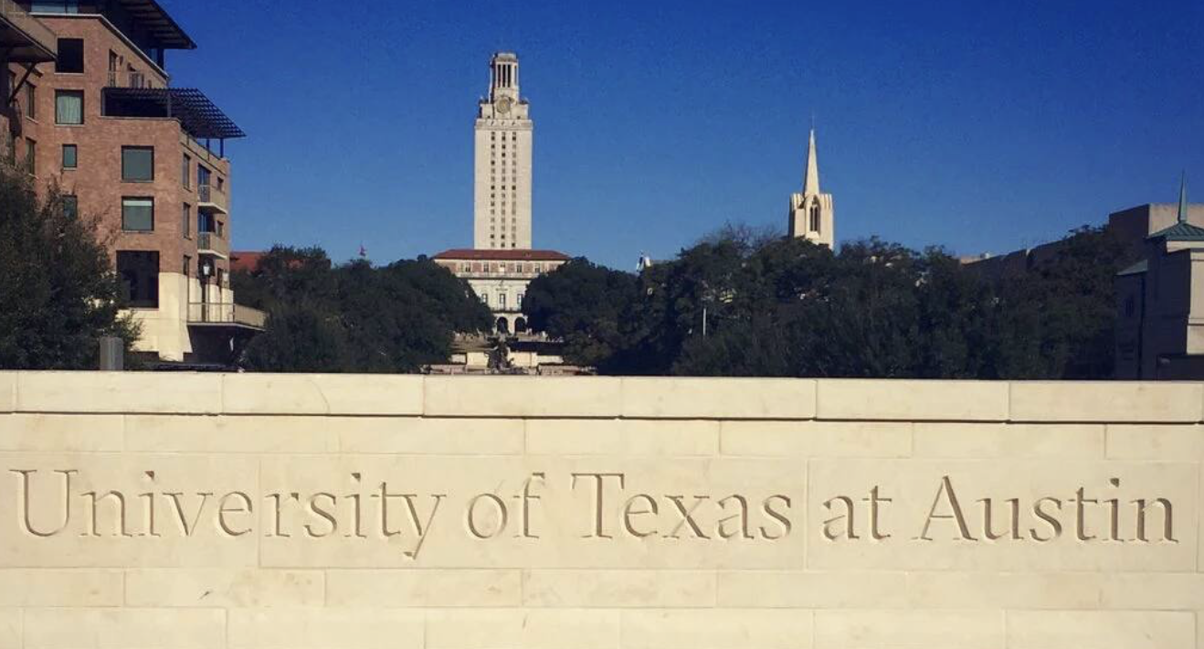 The University of Texas at Austin sign with a view of the university campus, including a tall building and a church steeple, under a clear blue sky.
