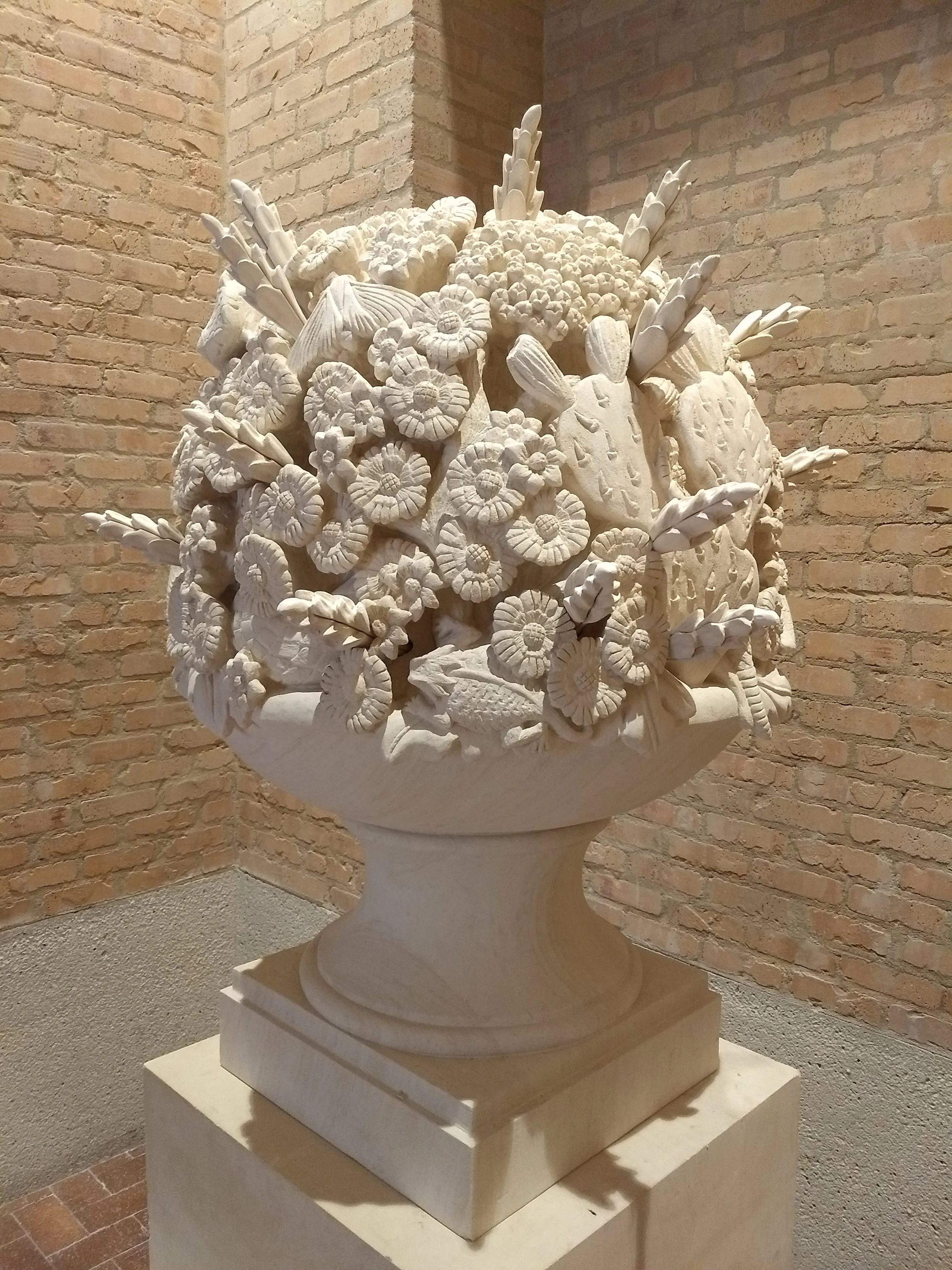 White marble sculpture of a classical urn filled with flowers and foliage, placed on a pedestal against a brick wall.