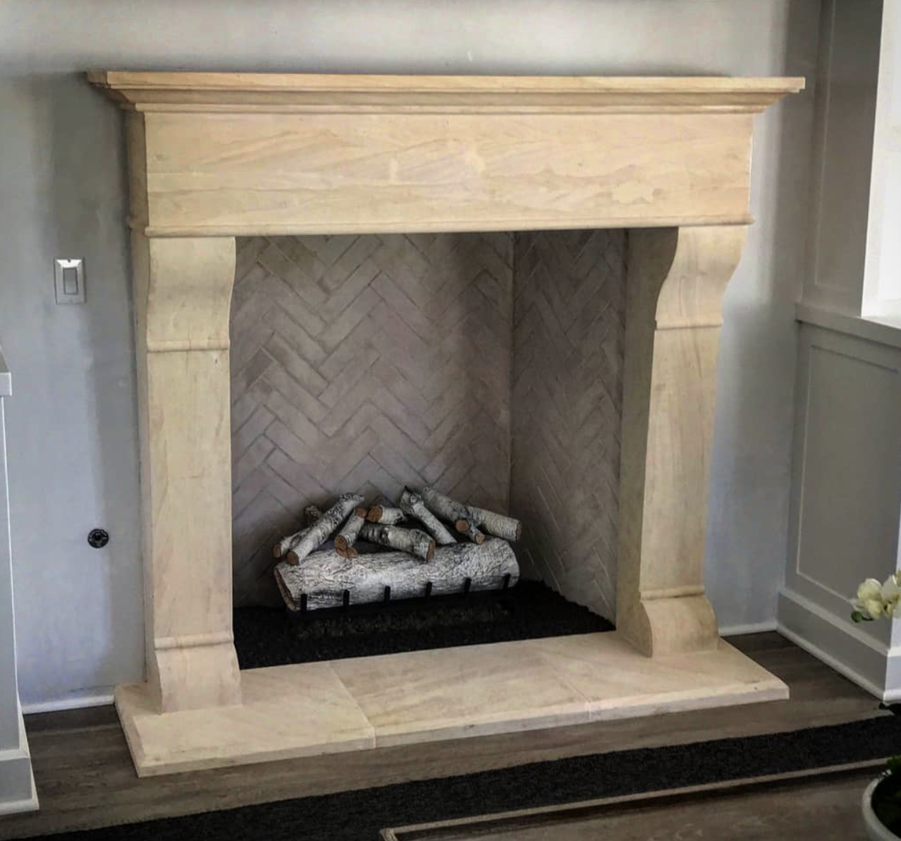 Empty fireplace with a beige stone mantel and interior lined with herringbone brick pattern; fake logs inside, set on a dark hearth in a living room.