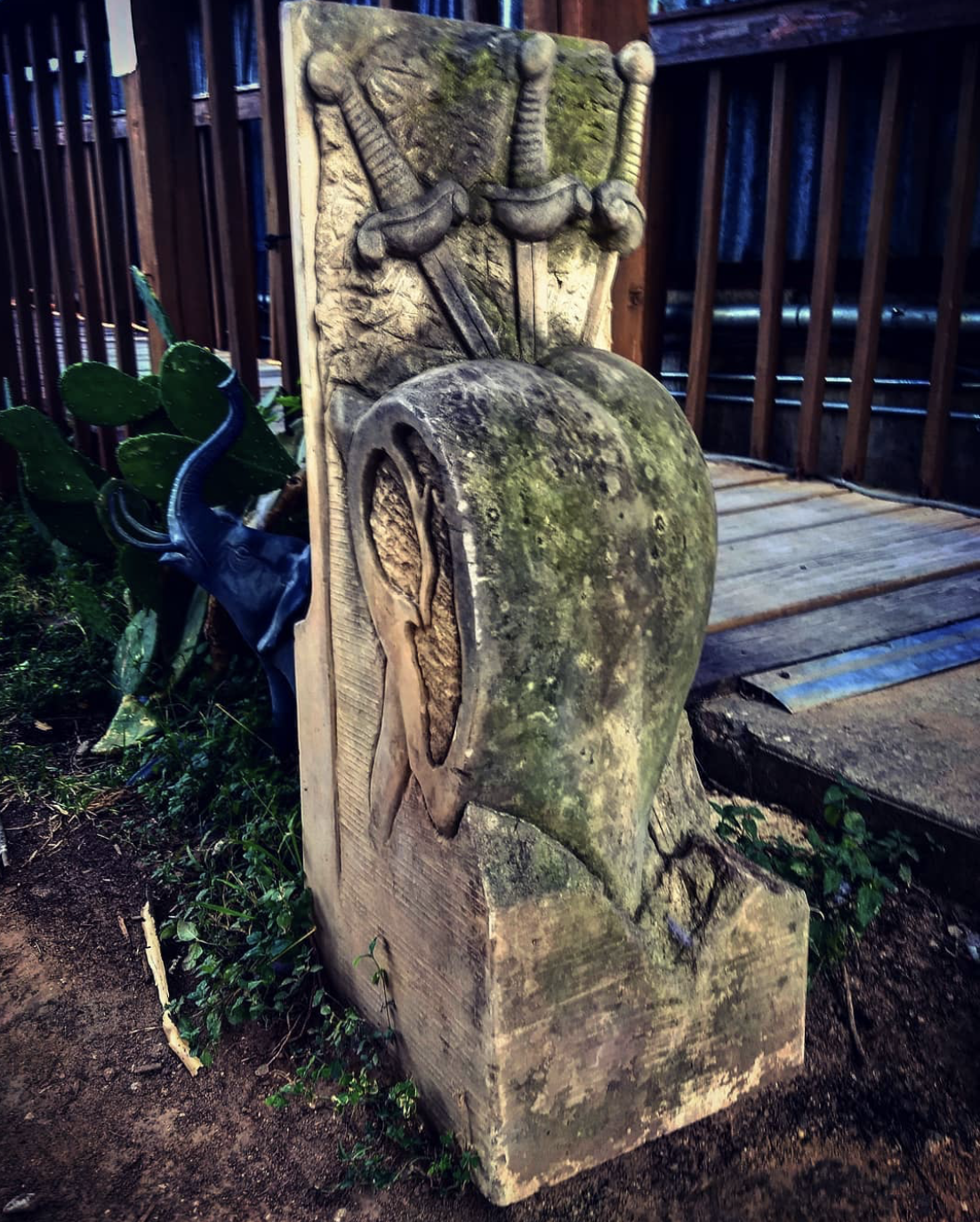 Carved wooden mailbox with decorative trim, moss and weathering, next to a cactus plant, on a dirt ground near a wooden deck.