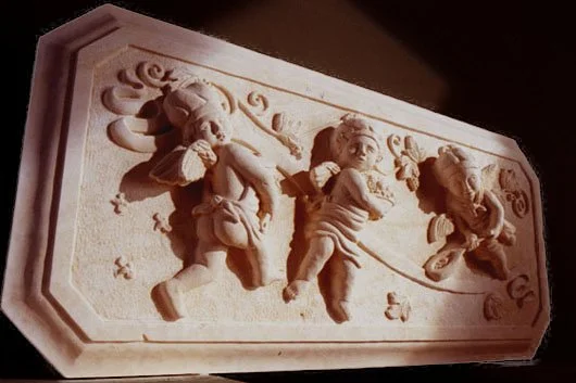 Carved stone relief of three cherubs carrying cooking tools with decorative scrollwork.