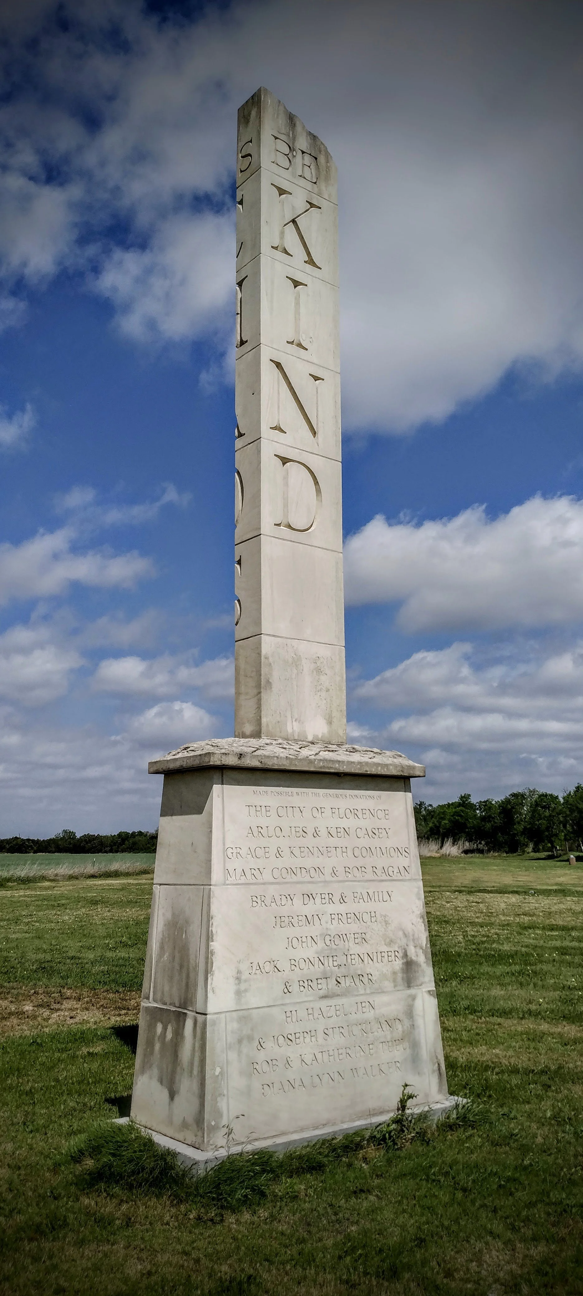 A tall white stone monument with the words "BE KIND" vertically carved into it, located in Florence, TX.  The base has inscriptions of donations from various individuals and organizations.