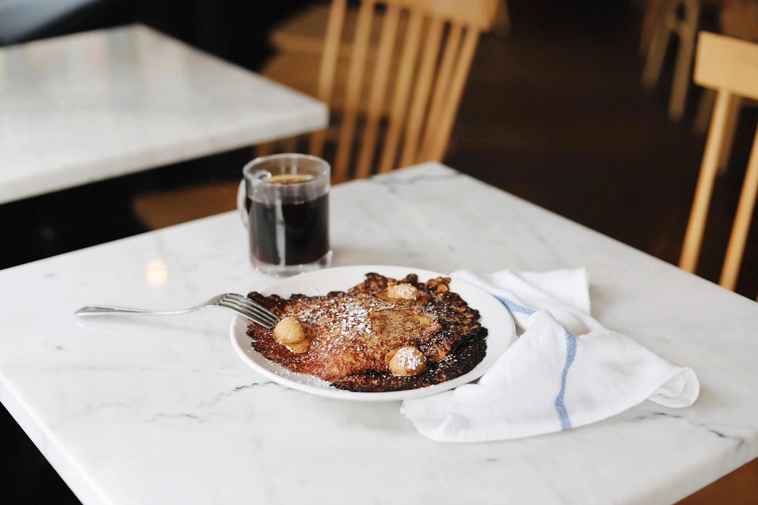 A plate of oatmeal griddle cakes sprinkled with powdered sugar and topped with dollops of butter, placed on a white marble table. A fork rests on the plate, and there is a cup of black coffee.