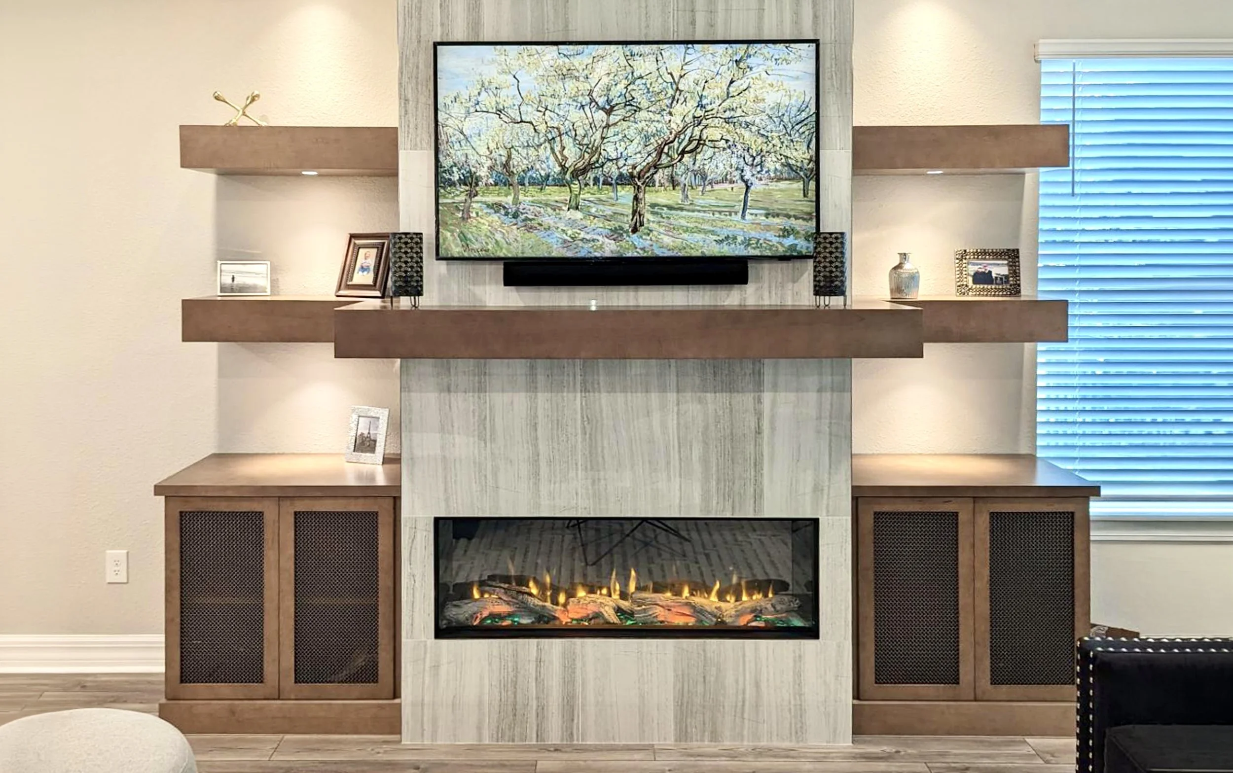 Living room fireplace with a large flat-screen TV above it, surrounded by a wooden and marble mantle, decorated with small framed photos and a vase, with a window with blinds on the right side.